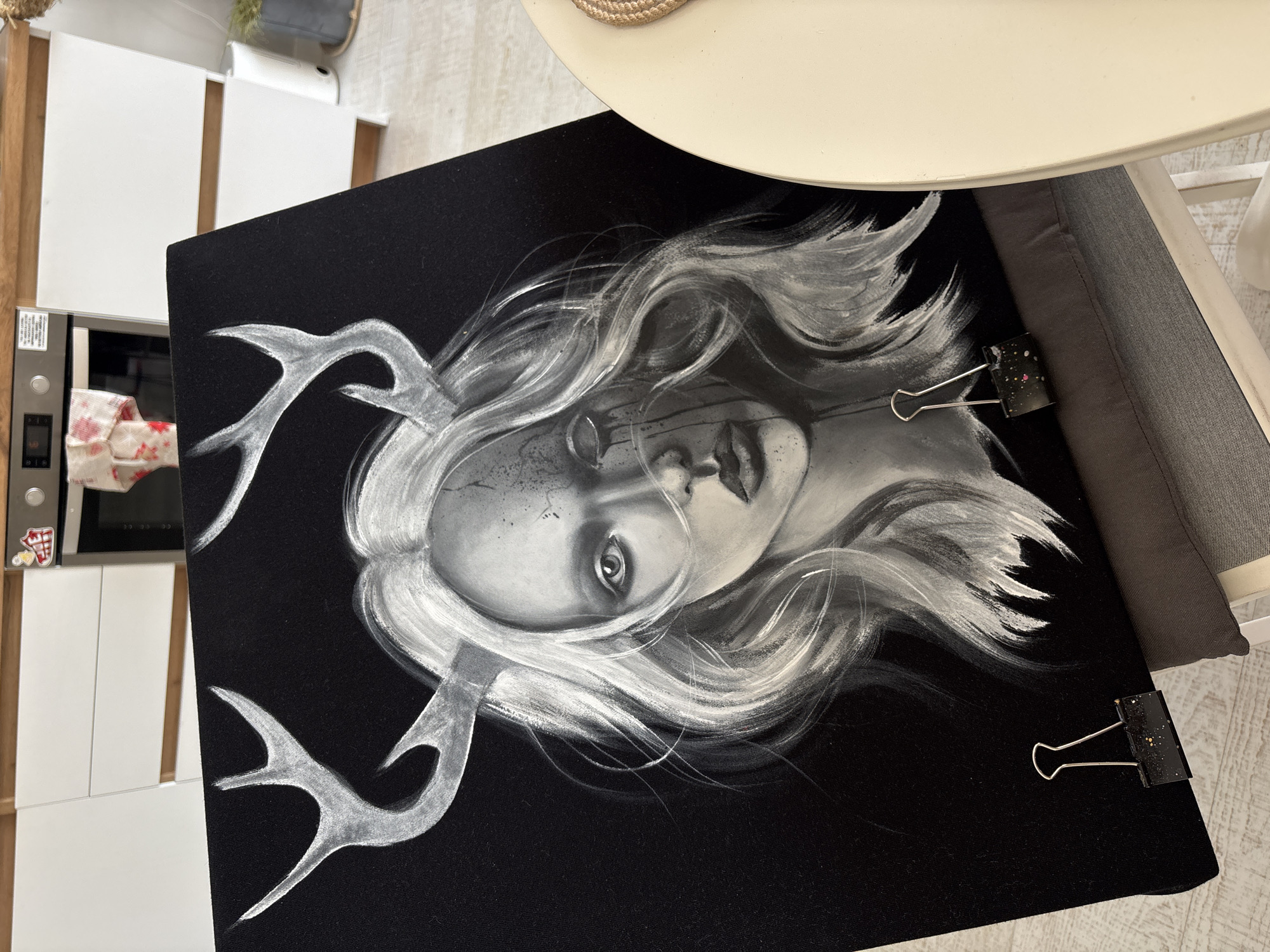 The image shows a close-up of a black canvas with a charcoal or graphite drawing of a pale-skinned woman with white hair and antlers. The drawing is secured with two black binder clips on the right edge. The background includes a white kitchen cabinet, a microwave oven, a round table with a white surface and natural fiber texture, and a gray cushion. The location is unknown but is not necessarily in Krasnoyarsk, Russia.