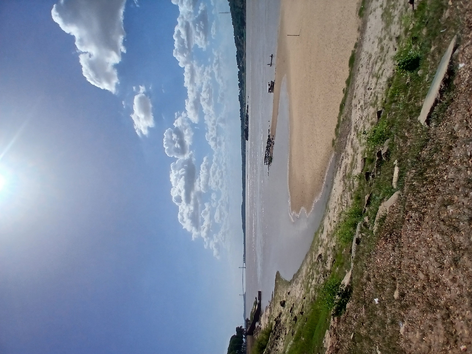 The image presents a view of the Orinoco River in Ciudad Bolivar, Venezuela, under a bright, partly cloudy sky. A sandy beach runs alongside the river's edge, transitioning into a grassy and rocky shoreline. The river contains several boats, while a distant bridge spans its width. The foreground shows a dirt and rock-covered area with sparse vegetation. The sun shines brightly in the upper left corner of the frame.