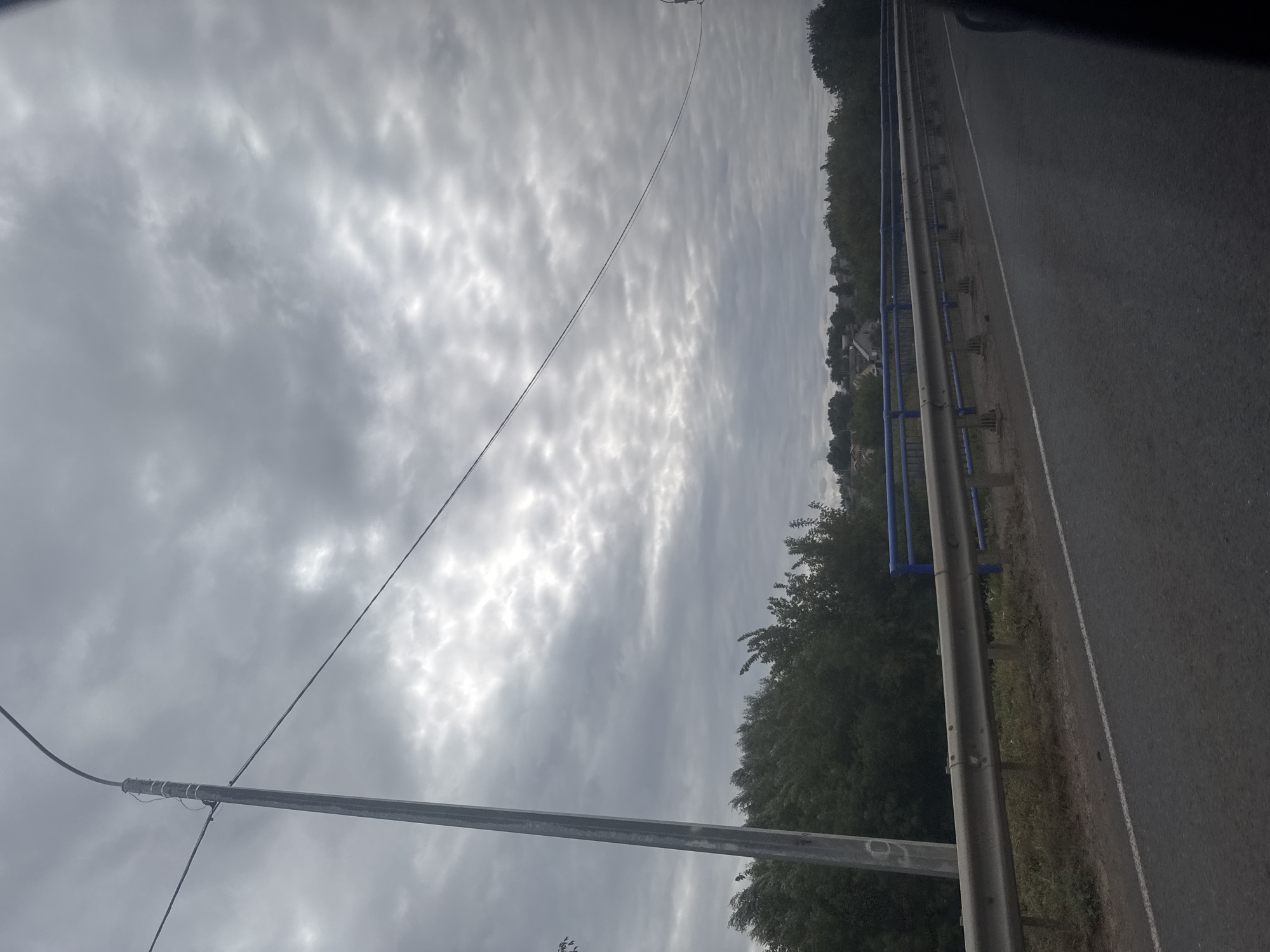 The image depicts a cloudy sky over a rural landscape. In the foreground, a paved road runs alongside a metal guardrail. Beyond the guardrail, a line of trees forms a dense border, with some houses visible through the foliage in the distance. A tall, gray pole, likely a lamppost, stands on the left side of the road, with wires stretching across the sky. The overall scene suggests a somewhat overcast day in a sparsely populated area. The location is identified as Novosergiyevka, Russia.