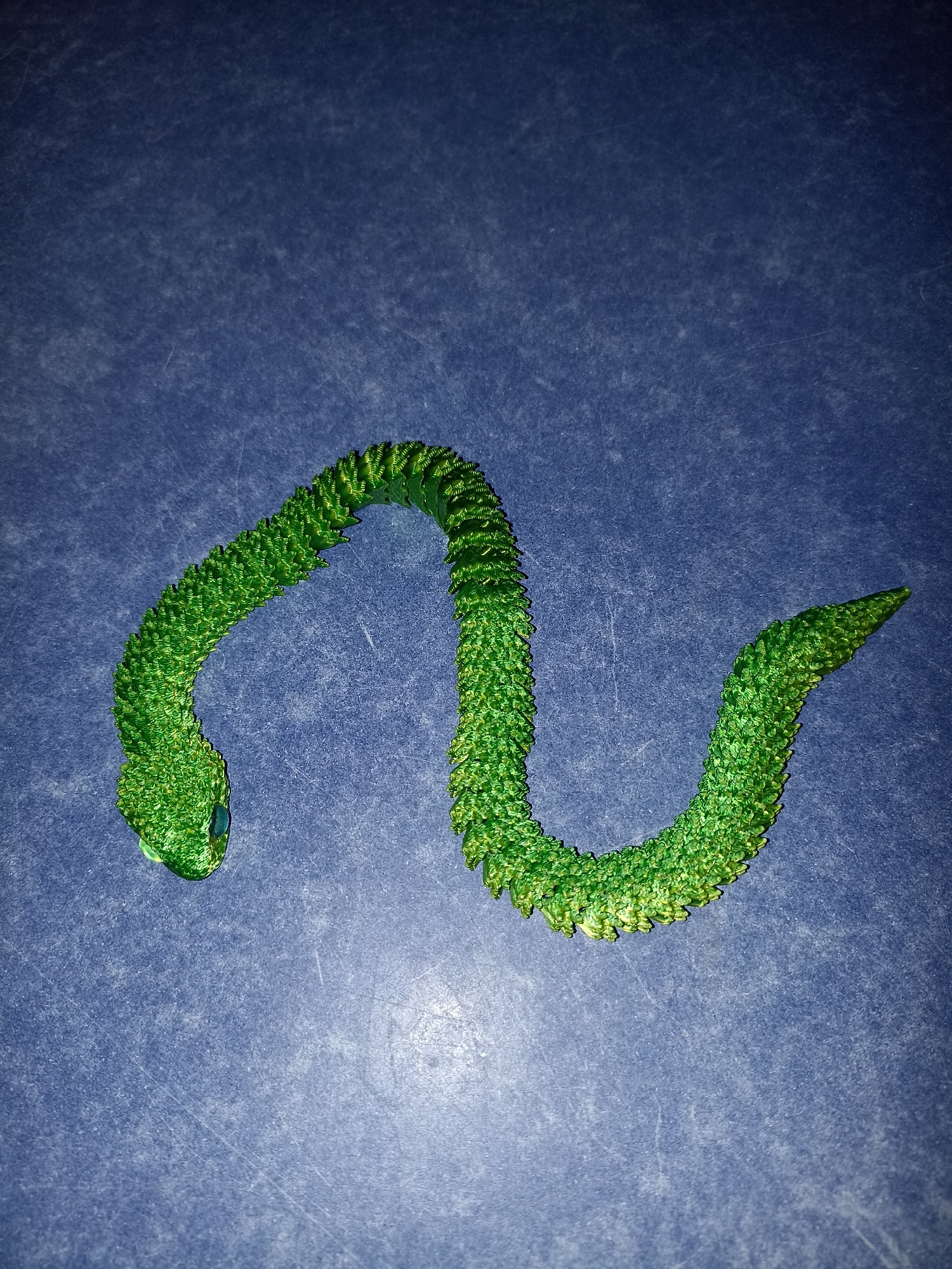 A green, segmented, toy snake is coiled in a 'W' shape on a dark blue, textured surface. The snake has small blue eyes and appears to be made of plastic. The surface has visible scratches and dust. The scene is lit from above, casting subtle shadows. The location is not discernible from the image.