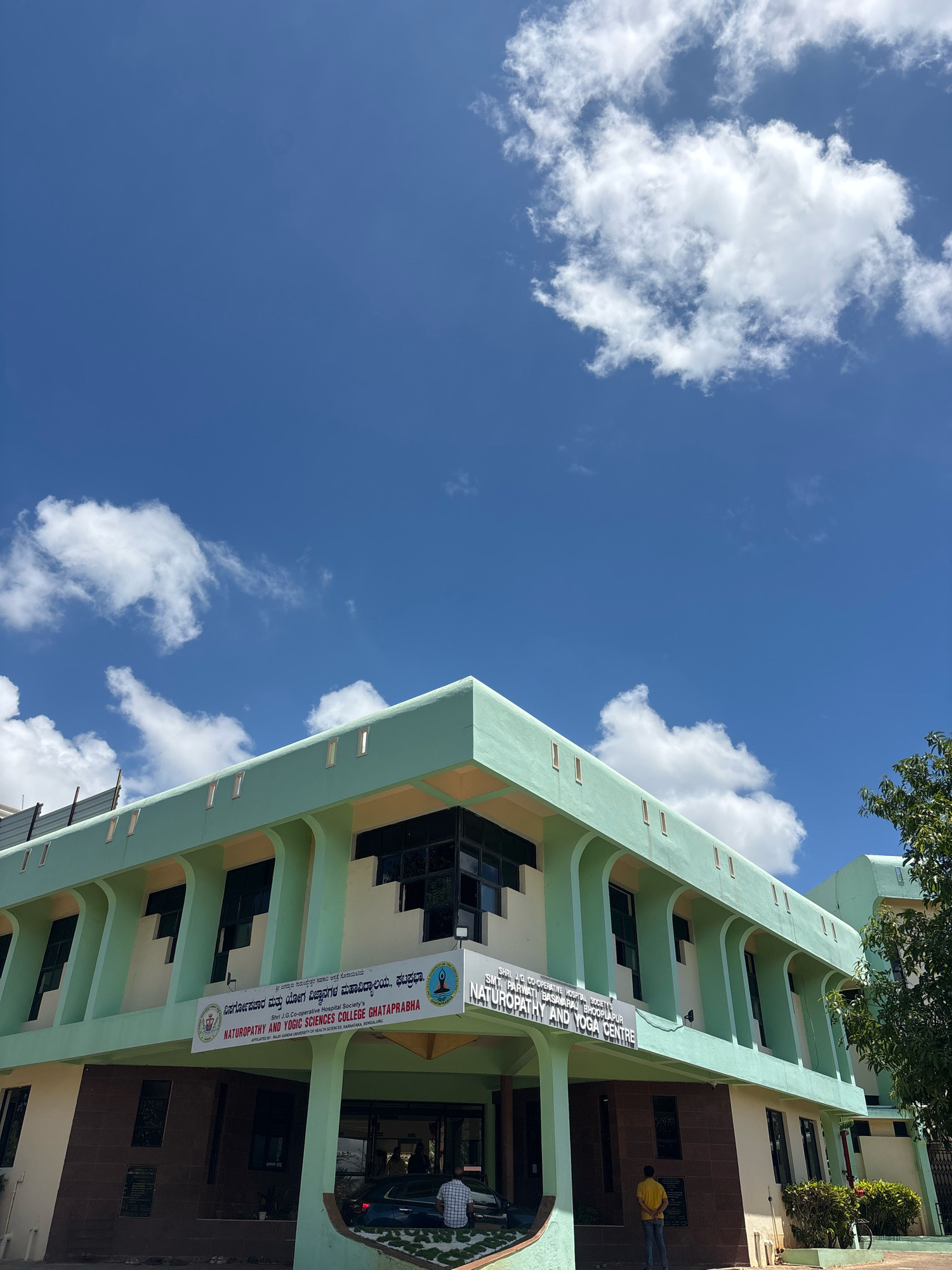 The image shows the exterior of a building, identified by signage as "Shri J.G. Co-operative Hospital Society's Smt. Parwati Basavaraj Ghataprabha Naturopathy and Yoga Centre." The building is painted in a light green and cream color scheme, with multiple floors and large windows. A clear blue sky with scattered white clouds is visible above the building. The location is stated as Jhangatihala, India. In front of the building, there is a paved area with some greenery and a parked car. A few people are visible near the entrance, one of whom is standing and looking at the building, and another is seated inside the car. The overall scene depicts a public institution on a bright, sunny day.