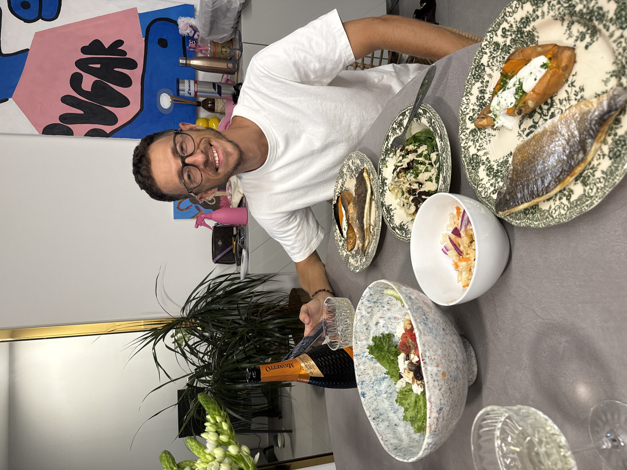 A man with glasses and a white t-shirt is smiling and seated at a table with food. He is holding a phone. The table is set with plates of food, including fish, sweet potato, and salad, as well as a bottle of Mionetto Prosecco and glasses of champagne. The room appears to be a dining area with a large plant and abstract art on the wall. The scene is likely in Tel-Aviv, Israel, as indicated by the man's presence in the city.