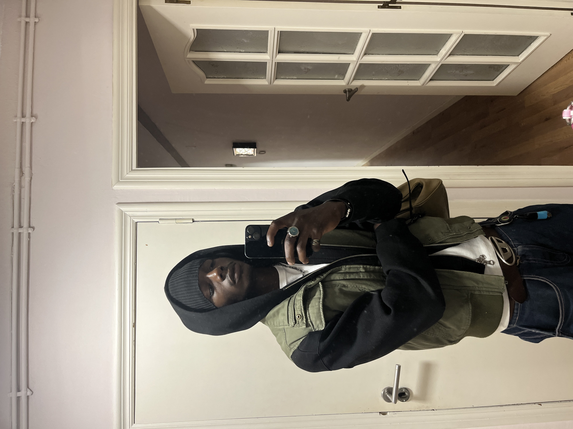 A medium shot depicts a man taking a selfie in a mirror. He is wearing a black hooded sweatshirt, a green jacket, blue jeans, and a grey beanie. The mirror reflects a hallway with a white door at the end. The location is inside an apartment or house, but the city of Barcelona cannot be confirmed from the visual information provided.