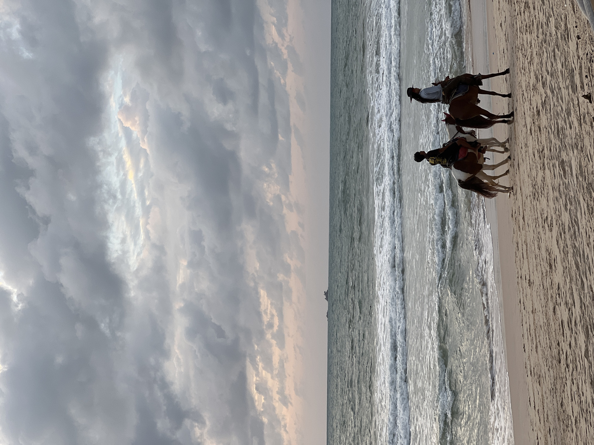 This image depicts two individuals on horseback riding along a sandy beach in Choeng Thale, Thailand. The sky above is filled with heavy, grey clouds, with some hints of light peeking through. The ocean is a murky green with visible waves breaking on the shore. In the distance, a small island or landmass is visible on the horizon. The sand is a light brown and shows some texture. The two riders are positioned side-by-side on the beach. The rider on the left is on a horse with brown and white markings, and is wearing a dark shirt. The rider on the right is on a brown horse and is wearing a light blue shirt and a dark hat. Both riders appear to be moving parallel to the water.