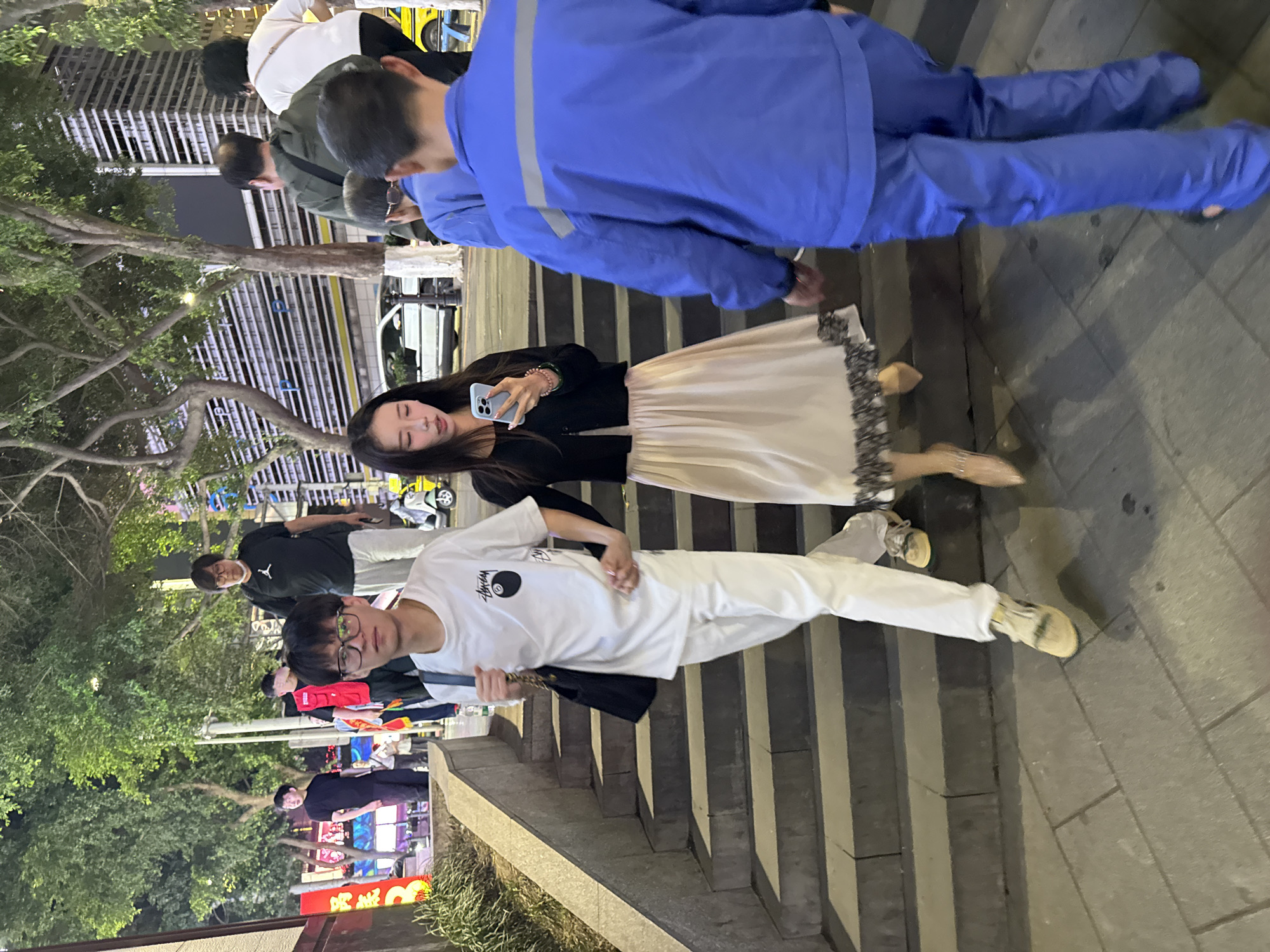 The image captures a street scene in the Yuzhong District of China, likely at night or during the evening, as indicated by the artificial lighting and visible store signs. A group of people are present, some walking on a set of concrete stairs that appear to be leading up from the street level.
A young woman is holding up a smartphone, possibly taking a photo or video. She is wearing a dark jacket and a light-colored skirt. Next to her is a young man wearing glasses, a white t-shirt with a graphic on it, and white pants, walking on the stairs. A man in a blue work suit is walking in the opposite direction.
Other people are seen in the background, walking or standing nearby, including a person wearing a red item of clothing. Storefronts with illuminated signage are visible, along with trees and other urban vegetation. The environment suggests a busy urban area with both pedestrian and vehicular traffic.