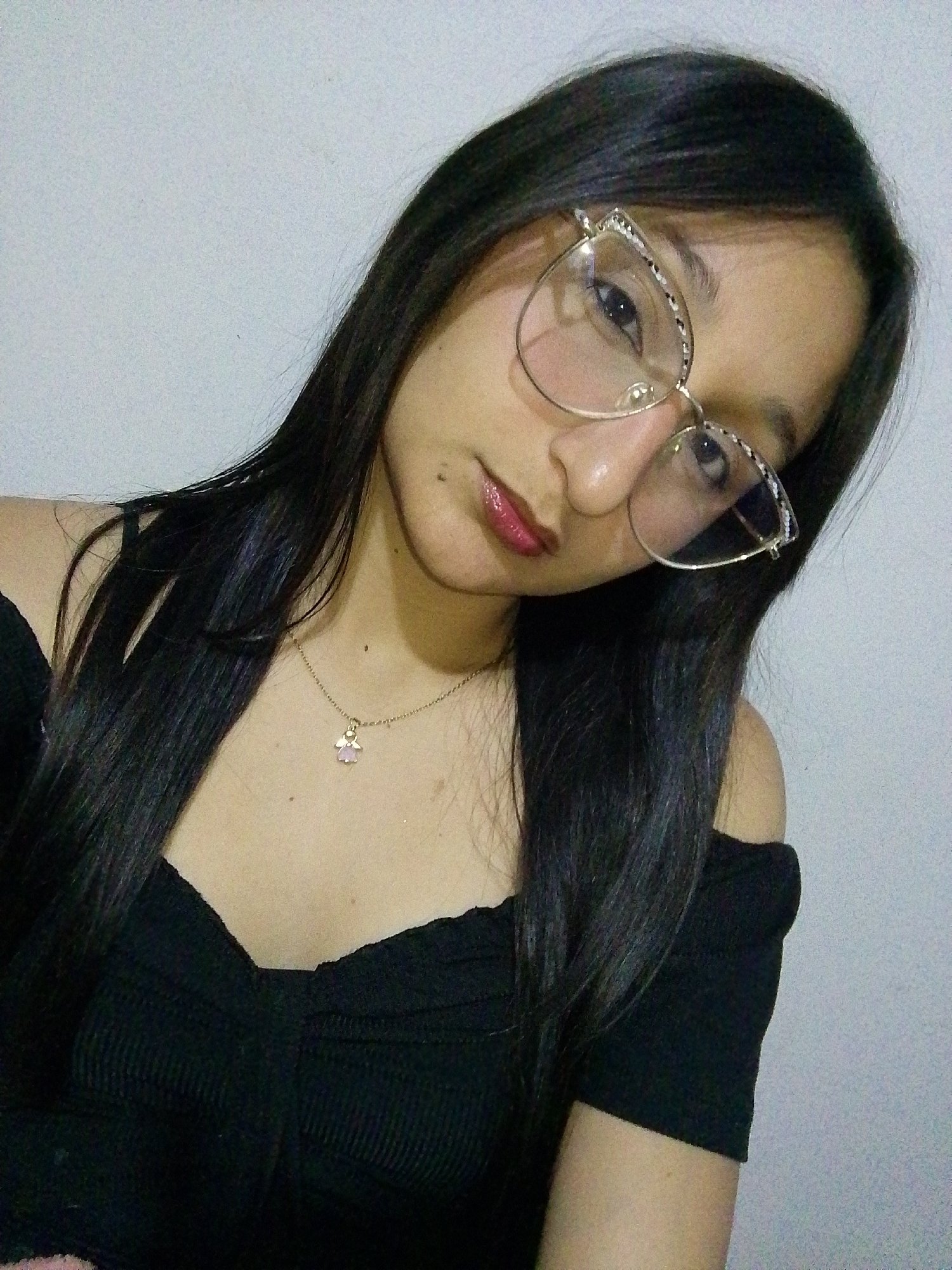 A woman poses indoors against a neutral background, slightly turned and looking down into the camera. She wears a black off-the-shoulder top and a delicate gold necklace with a pink pendant. Her dark, straight hair falls past her shoulders. She is wearing cat-eye glasses with gold frames. There are no other people, subjects, objects, actions, or interactions apparent in the photo. No location in Juan Griego, Venezuela can be confirmed based on the provided image.
