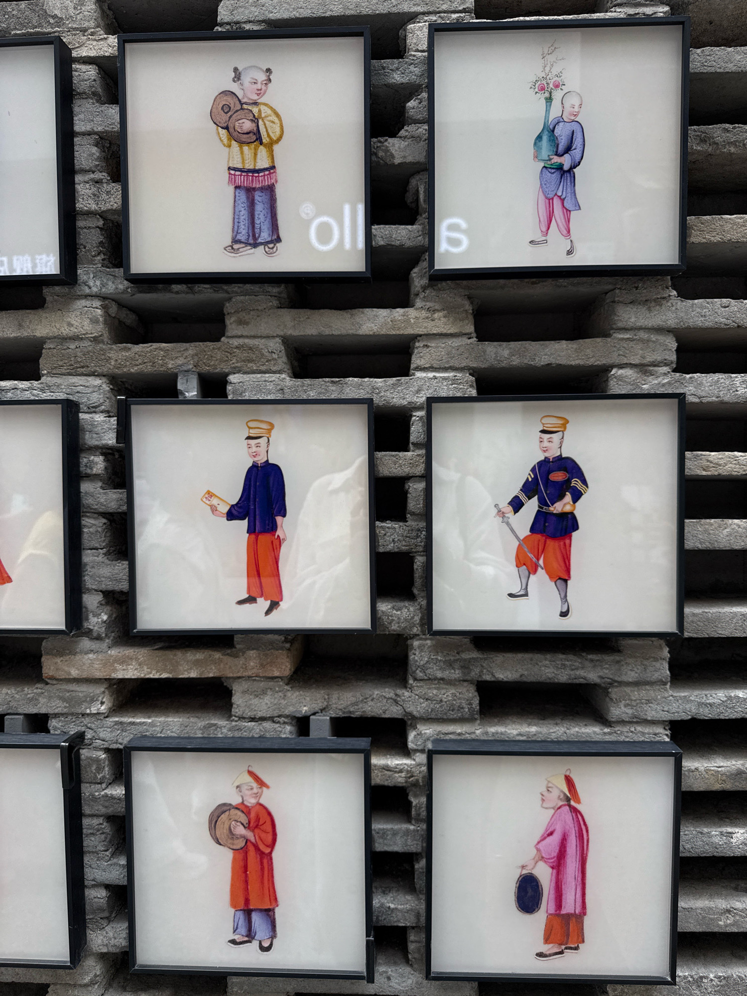 The image displays a wall constructed from rough, grey, rectangular stones arranged in a staggered pattern with horizontal openings. Mounted on the wall are several framed artworks, each featuring a painting of a person.
In the top row, the leftmost frame is partially visible and appears to be a blank white rectangle with some faint characters. The next frame to the right shows a person with short hair holding two round, brown objects in their hands. They are wearing a yellow robe with blue and pink trim and blue pants. To the right of this is another frame with a person holding a blue vase filled with flowers. This person is wearing a blue robe and pink pants.
The middle row features frames with people in more formal attire. The leftmost visible frame in this row shows a person wearing a blue jacket with gold trim, a yellow hat, and orange pants, holding a small rectangular object. The frame to its right depicts a person in a blue military-style uniform with gold epaulets and a yellow hat, holding a sword. They are also wearing orange pants.
The bottom row contains two frames, each showcasing a person holding a circular object. The leftmost frame in this row shows a person in a bright red robe and blue pants, holding a brown circular object. The frame to the right shows a person in a pink robe and orange pants, holding a dark blue circular object.
The background appears to be an outdoor setting, given the stone wall construction and the natural lighting. The artworks are square-shaped and uniformly framed in black. There are also some faint white characters visible on the wall next to the top row artworks, suggesting possible signage or decorative elements. The location is identified as Nanshan District, China.