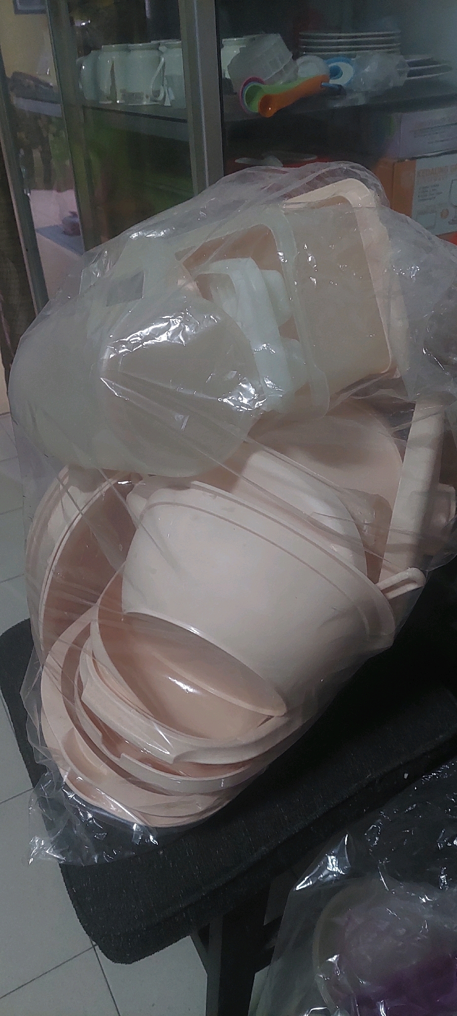 A pile of nested, light-pink plastic bowls is wrapped in a clear plastic bag, creating a bulky shape. The bowls have a subtle ribbed pattern on their outer surfaces. The bag is placed on a dark, textured surface that appears to be a cushion or upholstered seat, which is supported by dark legs. In the background, a glass-fronted cabinet is visible, containing stacks of white dishes, some clear plastic containers, and a set of colorful measuring spoons with orange, green, and pink scoops. The cabinet is lit from within, casting a soft glow on the items inside. The overall scene suggests an indoor storage or retail environment.