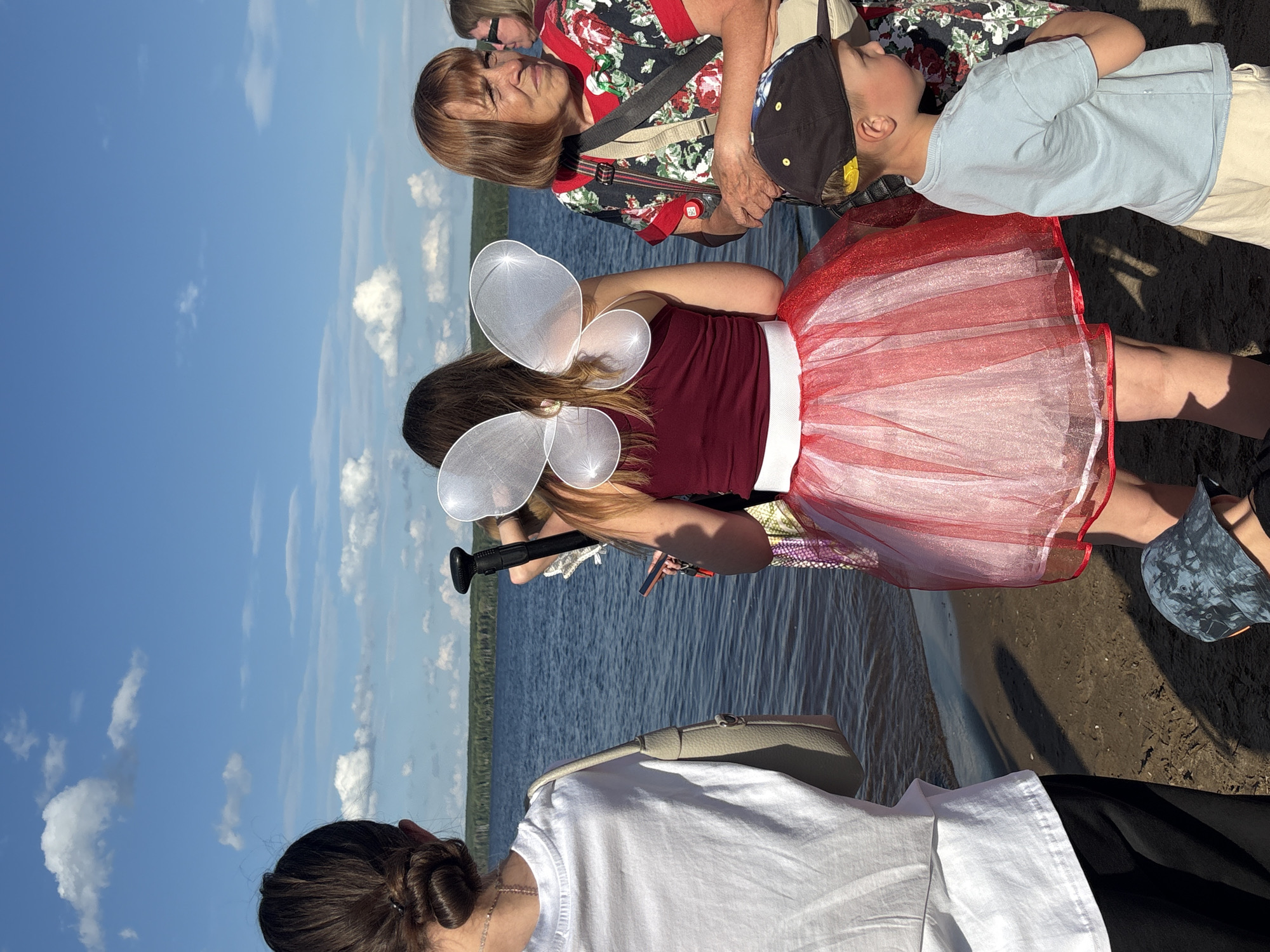 This is a photograph of a group of people standing near a body of water on a sunny day. The image is oriented vertically. In the foreground, a person wearing a white t-shirt and dark pants is partially visible, with their back to the camera. To their left, another person with their hair in a bun is also seen from behind.
In the middle ground, a person is dressed in a maroon top and a red and white sparkly tutu, with large white fairy wings attached to their back. They are holding a black cylindrical object. To their right, a young boy wearing a blue t-shirt and a dark baseball cap is looking upwards. Behind them, an older woman with short brown hair is wearing a red and black floral print top.
The setting appears to be a shoreline, with water and a grassy area visible in the background. The sky is bright blue with scattered white clouds, suggesting a pleasant day. The lighting indicates it is daytime, likely midday given the brightness. The overall mood seems relaxed and perhaps festive, given the attire of the person in the tutu. There is no visible text in the image.
