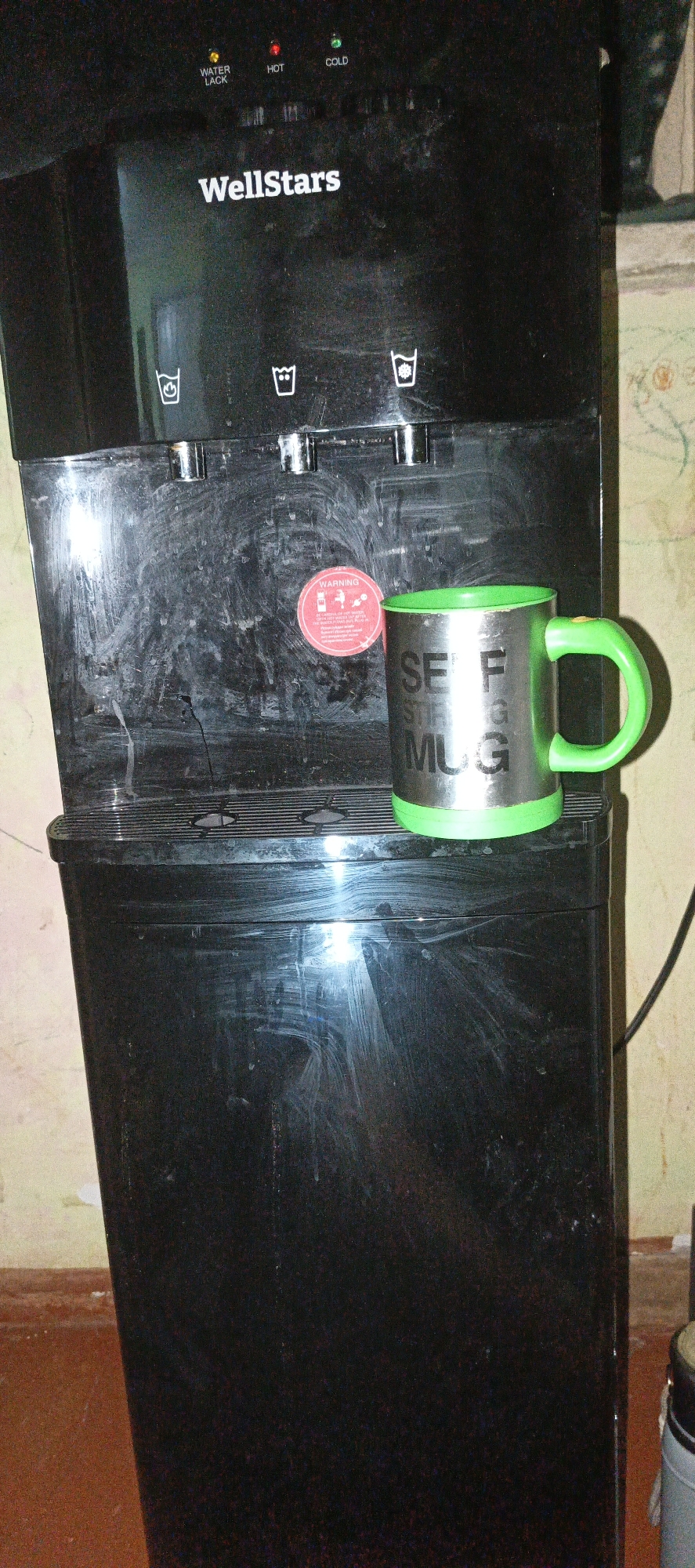 A stainless steel mug with a bright green handle and base sits on top of a black water cooler. The mug is inscribed with the words "SELF STIRRING MUG." The water cooler has three dispensing spigots, each with an icon above it. The left icon depicts a cup, the middle icon a beaker, and the right icon a cup. Above the spigots are indicator lights labeled "WATER LACK" (yellow), "HOT" (red), and "COLD" (green). The brand name "WellStars" is visible on the water cooler. A red circular sticker with "WARNING" and other text is affixed to the front of the water cooler. The water cooler is placed in an indoor environment with a cream-colored wall in the background and a reddish-brown floor. The water cooler appears to be dusty and smudged. No people are present in the image. The location is Aktash, Uzbekistan.