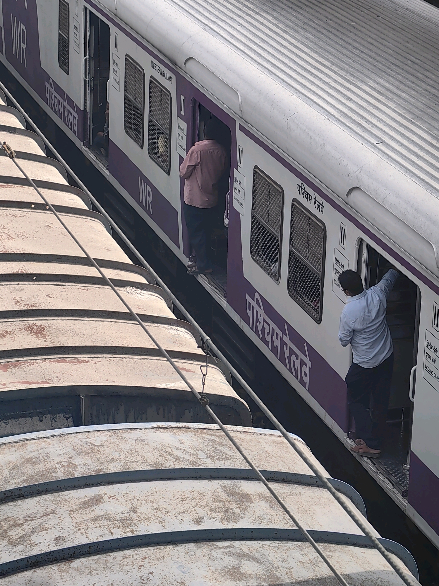 The image shows a train at a station in Mumbai, India. The train is predominantly white with purple accents. The text "WESTERN RAILWAY" and "WR" are visible on the train, along with "पश्चिम रेलवे" in Hindi. Several people are visible on or near the train. One person in a light blue shirt and dark pants is standing at the open doorway of a train car, appearing to be interacting with someone inside. Another person in a pink shirt and dark pants is inside the train car. The train is adjacent to other train cars, with their ribbed, metallic roofs visible in the foreground. The environment appears to be an outdoor train station setting with the characteristic Mumbai atmosphere.
