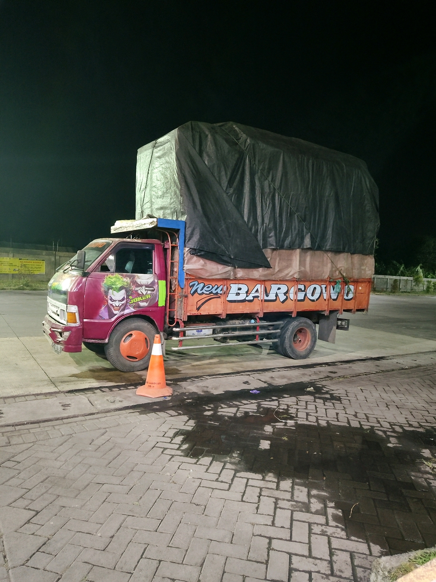 A maroon truck with a dark tarp covering its cargo is parked on a paved surface at night. The truck is decorated with images of the Joker on its side, with the word "JOKER" visible. The side of the truck also has the words "Neu Bargow" painted in white letters with a blue outline. An orange traffic cone is placed in front of the truck's front tire. The ground is wet and reflects the truck's lights. The background shows a fence and some trees. The image appears to be taken in Kedung Sukodani, Indonesia, given the information provided.