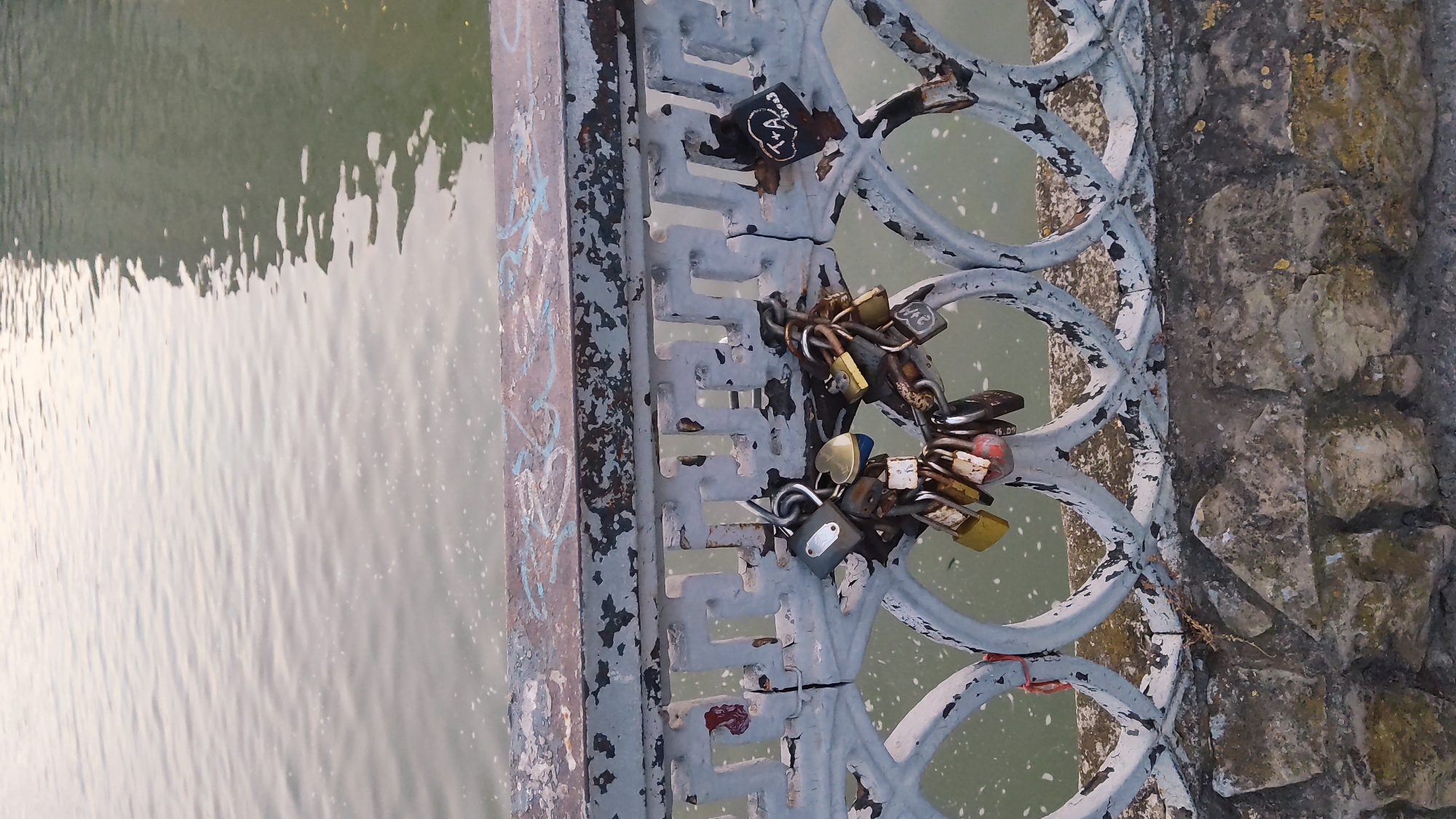 The image shows a close-up of a weathered, light blue painted metal railing on a bridge over a body of water. Numerous love locks are attached to the railing, forming a heart shape on the left side of the frame. The locks are of various sizes and colors, with some appearing older and more rusted than others. One black heart-shaped lock on the right side has white writing that appears to say "T+A 2003". Below the railing, a stone embankment is visible. The water is calm with ripples, reflecting a muted light. The scene is likely located in Krasnodar, Russia, a city known for its bridges adorned with love locks.