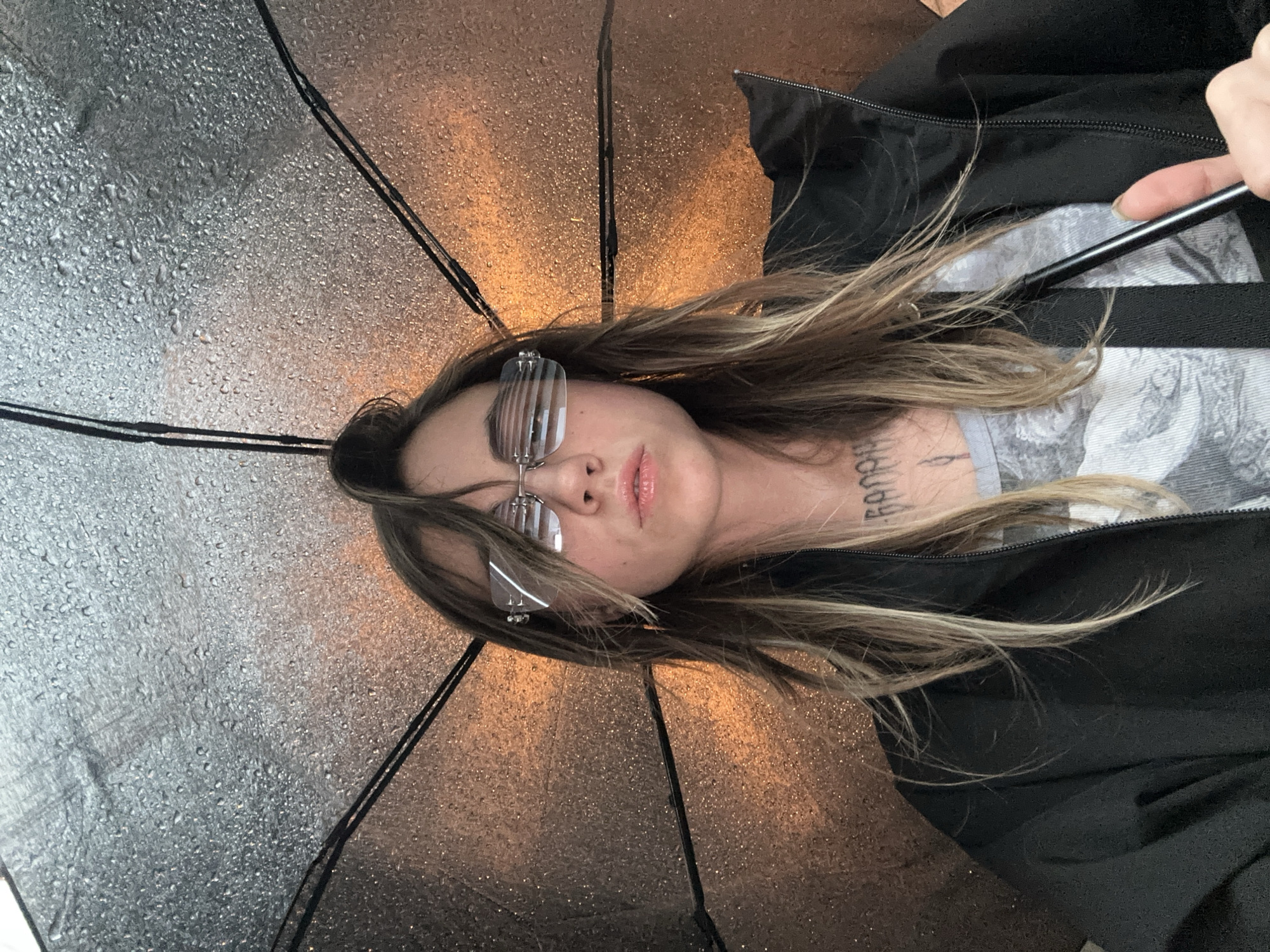 A close-up, over-the-shoulder shot shows a person holding an open umbrella indoors. The umbrella, made of a dark, translucent material, has numerous water droplets on its surface, suggesting it may have recently been used outdoors or is being used to collect water. The backdrop behind the umbrella is illuminated by a warm, orange light, creating a hazy and somewhat abstract background. The person is wearing a black jacket and glasses with a distinctive, horizontal slatted design. Their hair is partially pulled up, with strands falling around their face. A tattoo is visible on their neck. The image does not provide any specific details that would allow for a determination of location within Moscow, Russia.
