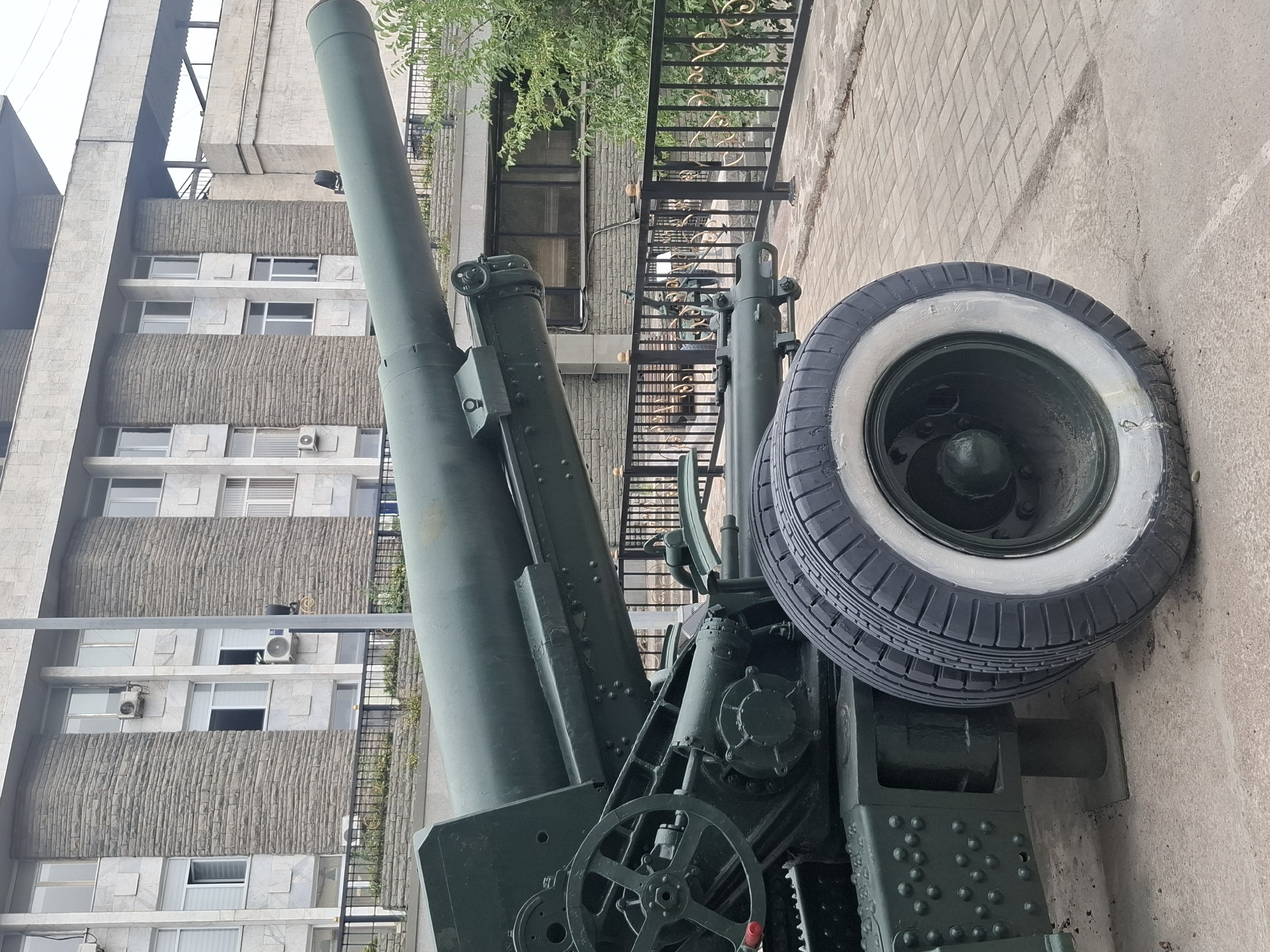An exterior medium shot shows a large, dark green artillery piece mounted on wheels. The cannon barrel is angled upwards, extending towards the right of the frame. The carriage features visible gears, a large spoked wheel on the left, and two tires with white rims in the foreground. The artillery piece is positioned on a paved walkway made of light-colored rectangular bricks.
Behind the artillery, a modern building with a stone facade and multiple windows is visible. A black metal fence with ornate gold detailing separates the walkway from the building's grounds. Green foliage can be seen above and to the right of the fence. The sky is overcast. The scene is devoid of any people or animals. The overall impression is one of a static display, likely a monument or historical exhibit, in an urban setting.
Based on visual cues such as architectural style and the artillery piece itself, this location is likely the Museum of Folk Musical Instruments named after Ykhlas in Almaty, Kazakhstan.