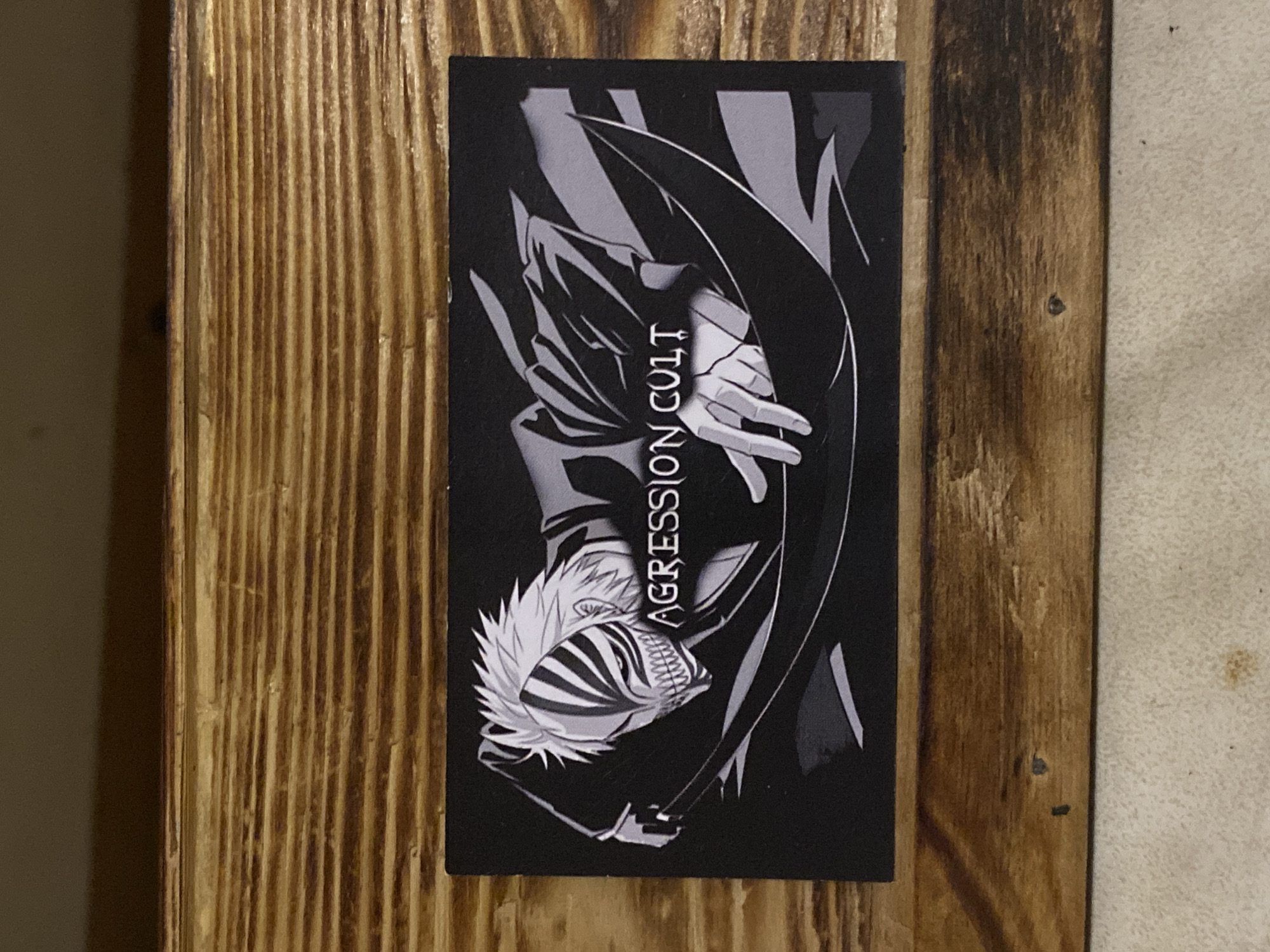 The image shows a black rectangular sticker with a monochromatic anime-style illustration of a character wearing a mask and a large, dark robe. The character has spiky white hair and a distinctive mask with black markings that cover the eyes and extend down the face, revealing sharp teeth. The text "AGRESSION CULT" is written in white, stylized font across the character's chest area. The sticker is affixed to a wooden surface with a visible wood grain pattern, suggesting it is a wooden box or furniture. The background behind the wooden surface is out of focus and appears to be a neutral-colored wall or surface. There is no discernible connection to the city of Bor, Russia, as there are no visual cues within the image that indicate this location.
