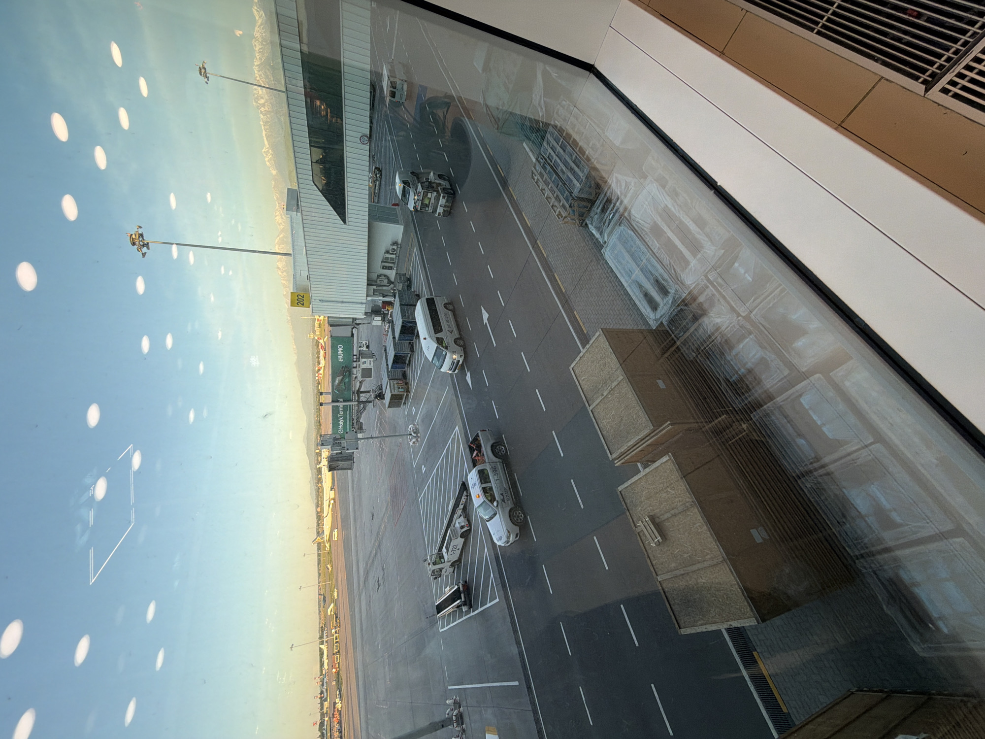 The image appears to be taken from inside a building, possibly an airport terminal, looking out onto a runway or tarmac area. The location is likely Almaty International Airport in Kazakhstan, given the partial signage of "Halyk Terminal" visible. Several ground vehicles, including vans and service vehicles, are present. The view is through a window, with reflections of the interior visible on the glass. The sky is clear with some atmospheric haze. Overall, it depicts a typical airport scene of ground operations and aircraft servicing.