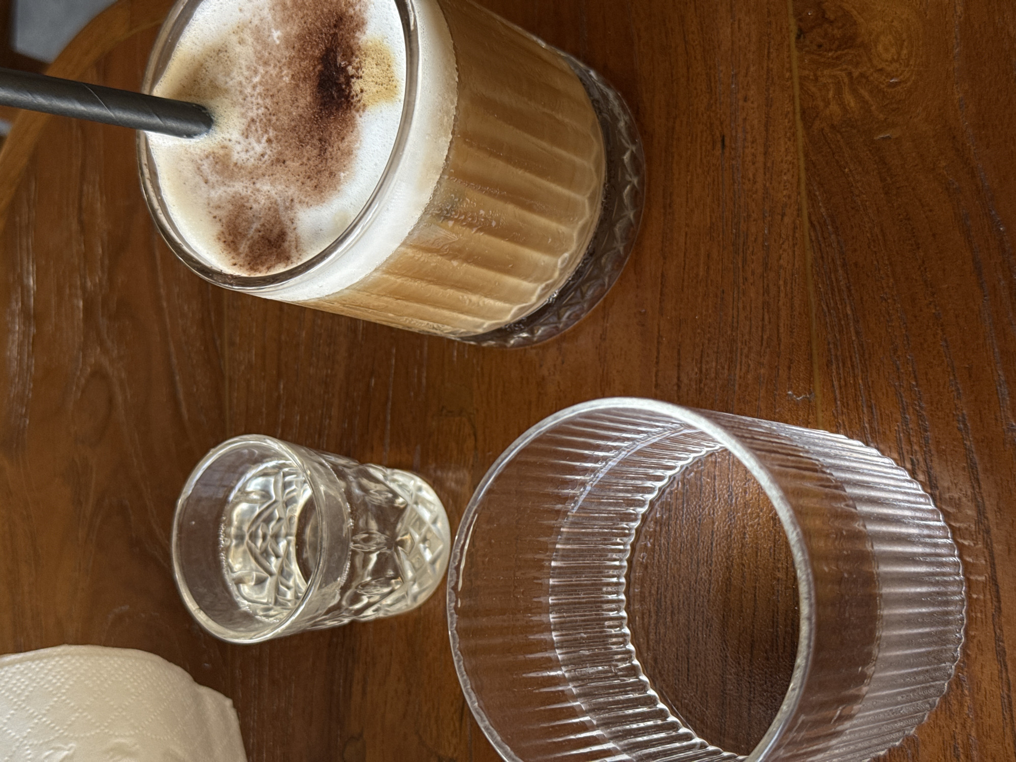An overhead, slightly angled shot captures a casual table setting on a warm-toned wooden surface. In the upper left, a clear glass filled with a coffee drink, likely a latte or cappuccino, takes prominence. The drink features distinct layers: a dark brown liquid base, topped with creamy white foam, and a dusting of dark brown powder, possibly cocoa or cinnamon, creating an artistic, swirled pattern on the surface. A dark grey or black paper straw is inserted into the coffee.
To the lower right of the coffee glass, two other clear glass items are visible. One is a small, empty, ornate glass with a faceted or diamond-cut pattern, positioned upright. Adjacent to it, lying on its side, is a larger, empty clear glass or bowl, distinguished by pronounced vertical ribbing around its exterior. A corner of a white, textured paper napkin or tissue is visible in the bottom left of the frame. The wooden table surface shows natural grain patterns and a rich, reddish-brown hue. No people or visible text are present in the image. The scene suggests a moment of relaxation, possibly at a cafe, restaurant, or home.