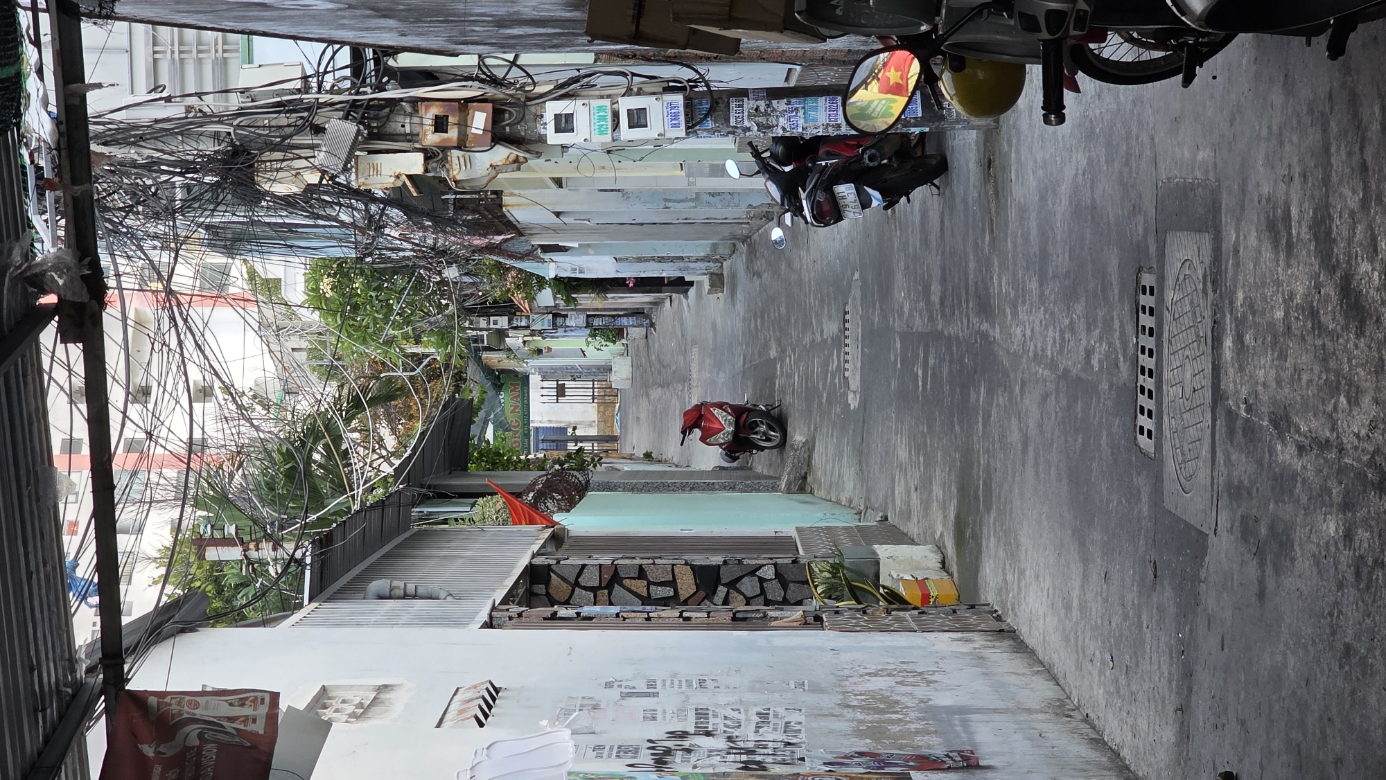 The image depicts a narrow alley in the city of Bac Nha Trang Ward, Vietnam. The alley is flanked by buildings with visible electrical meters and a dense network of tangled overhead electrical wires. The alley floor is concrete, showing some drainage covers. Several parked motorbikes are visible along the alleyway, one prominently red, and two others closer to the viewpoint. Signs and posters are pasted on the lower portions of the buildings, and the overall impression is of a densely populated, urban environment.