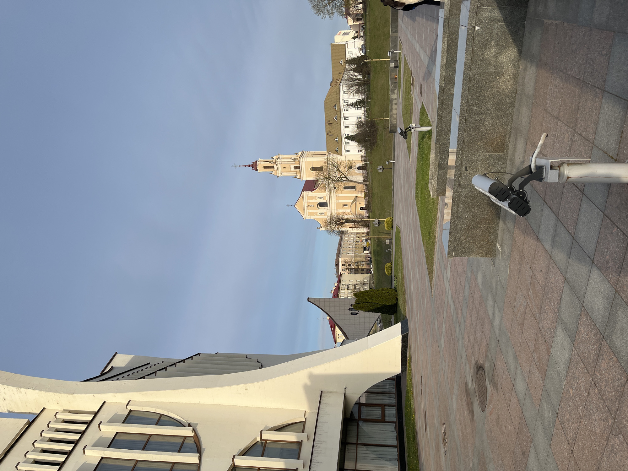The image shows a cityscape in an unknown city. In the background, there's a tall, yellow church-like building with a red roof and spire. Adjacent to it is a white, multi-story building. Closer to the foreground is a modern-looking, white building with curved walls and arched windows. The ground is covered with a paved area, patterned in red and gray. A security camera is mounted on a pole to the right of the frame. The sky is a clear, light blue. No people are clearly visible in the image.