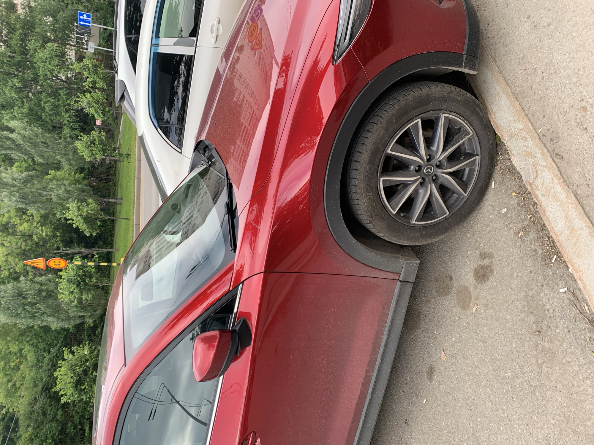 The image shows a red car parked on a street in Perm, Russia. The car is partially covered in dust. In the background, there are trees, a traffic sign indicating a speed limit of 40 km/h, and another car. The street appears to be made of asphalt, with some dirt and debris on the side. The overall scene is typical of an urban environment.