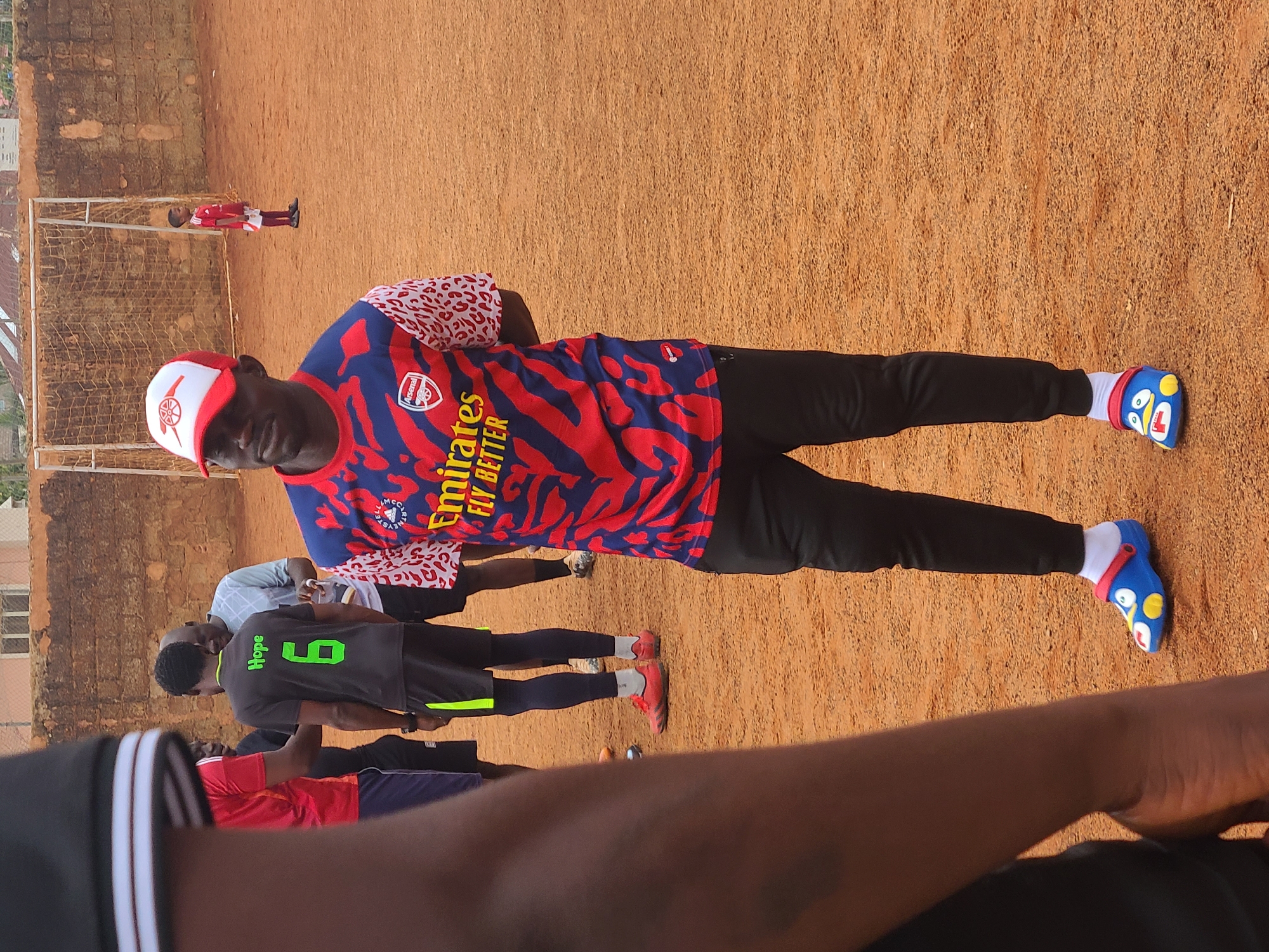 The image shows a group of people on a dirt field in Barakin Lerawi, Nigeria.
One man stands out, wearing an Arsenal soccer jersey, black pants, and red and white cap. He also wears white socks and blue and yellow clogs. He is standing with his arms spread.
In the background, another person is standing near a soccer goal. Another person is wearing a black jersey with the name "Hope" and the number "6" on the back. Other individuals are present in the scene, standing around.
The background includes a low brick wall. The ground appears to be hard-packed dirt, typical of a soccer field in the area.