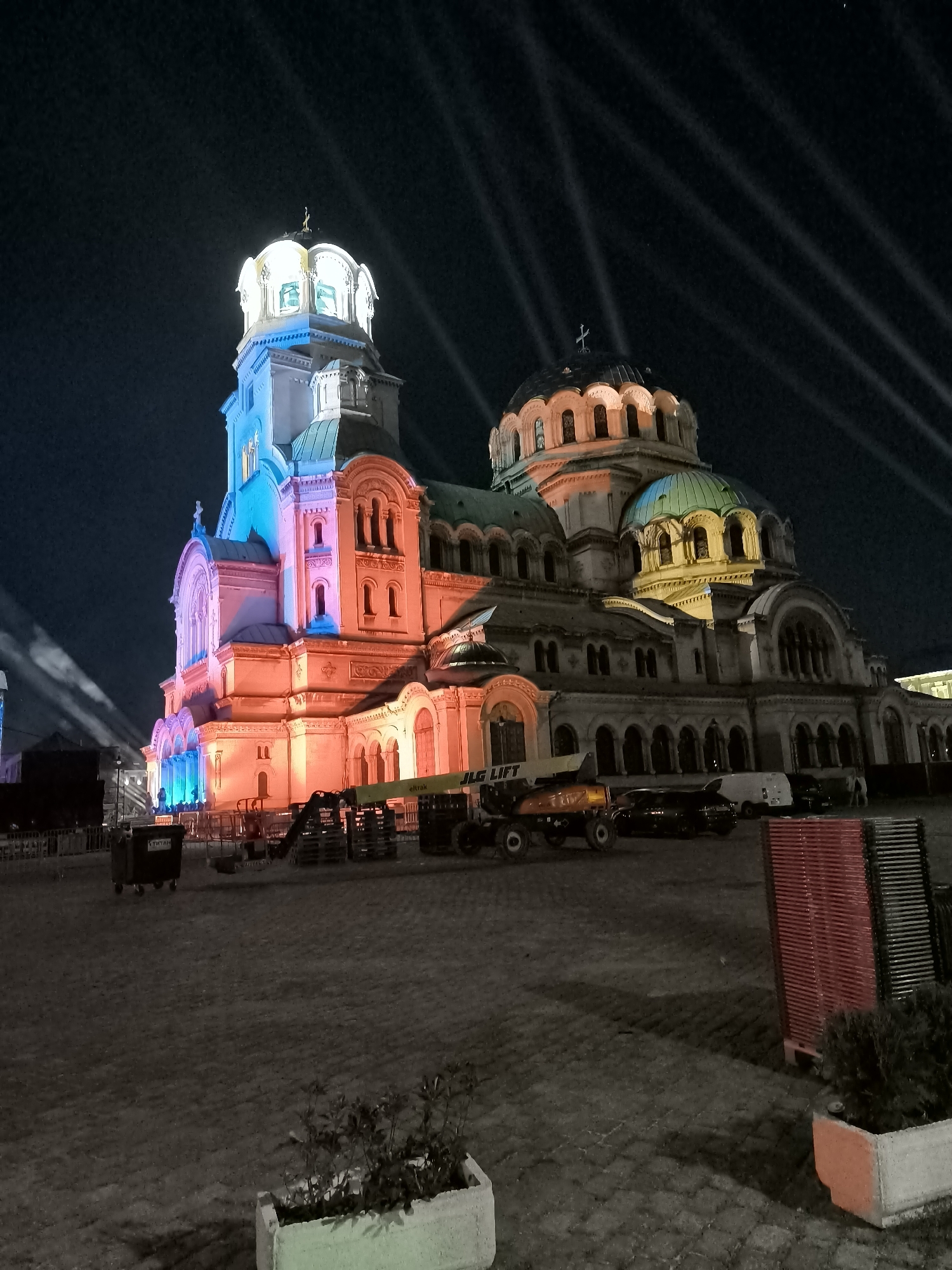 The image depicts the St. Alexander Nevsky Cathedral in Sofia, Bulgaria, illuminated at night by an array of colorful lights and spotlights. The cathedral's iconic domes and spires are prominently featured against a dark sky streaked with light beams. In the foreground, the paved plaza in front of the cathedral is visible, with several construction or event-related items scattered about. These include a large dark dumpster, several pieces of industrial equipment such as a scissor lift with "JLG LIFT" visible on its side and what appears to be a forklift, and stacks of red and black chairs. Several vehicles, including dark sedans and a white van, are parked behind the equipment. The overall scene suggests preparations for or the aftermath of an outdoor event or exhibition at this prominent Sofia landmark.