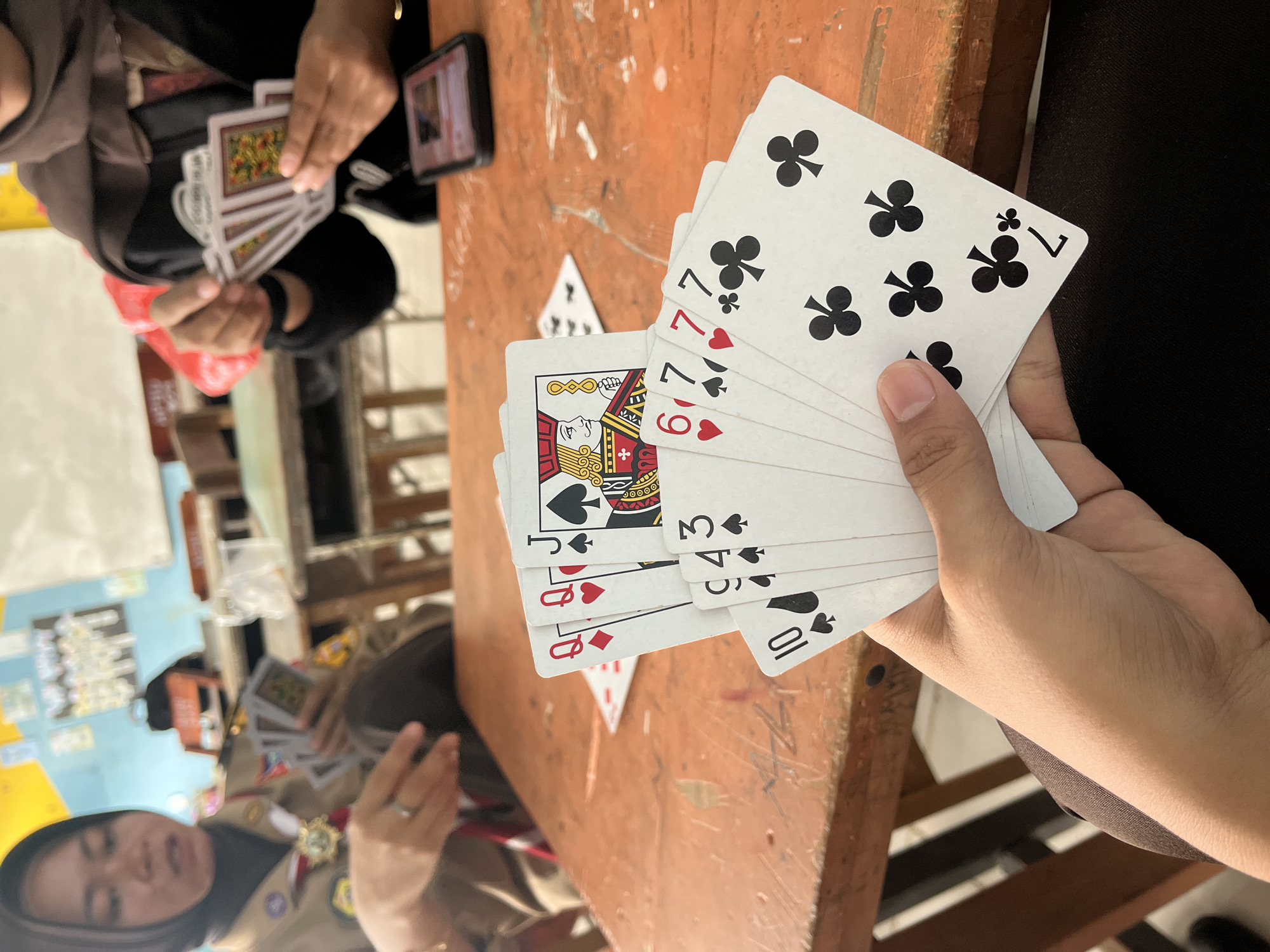 In Pompanua, Indonesia, two young women are playing a card game at a wooden table. The woman on the left, wearing a brown and beige uniform with a hijab, holds several cards in her hand, with her left hand resting on the table. The woman on the right, also wearing a hijab, is in the process of placing cards on the table, with a smartphone visible in front of her. The table is an orange-brown wood with visible grain and some scratches. Scattered on the table are other cards, with visible suits of diamonds, hearts, spades, and clubs. The background shows a wall with some posters or decorations, and further tables and chairs, suggesting a classroom or a communal area. The lighting is bright, casting shadows on the table.