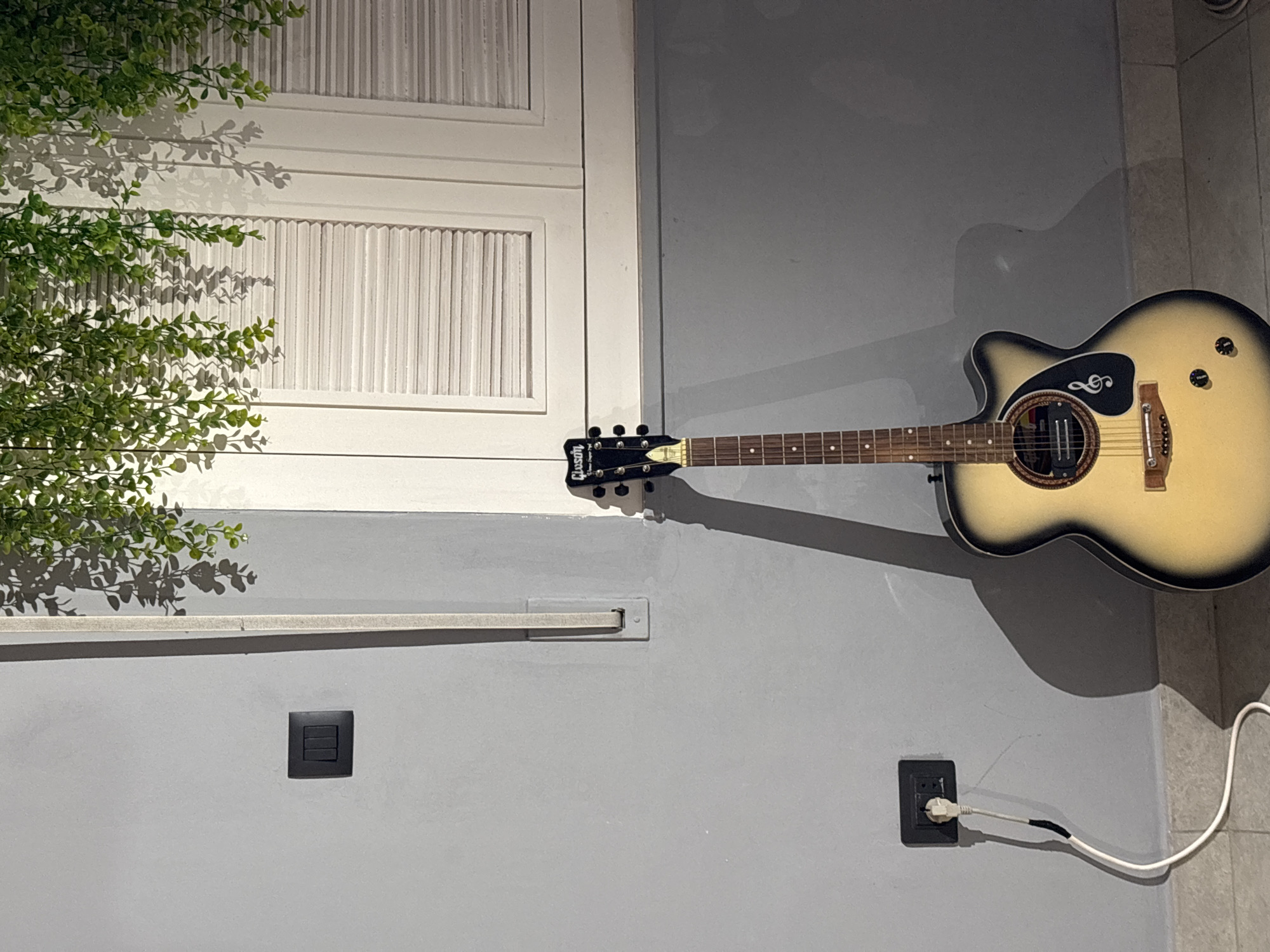 In the city of Sweileh, Jordan, an acoustic guitar stands upright against a light gray wall. The guitar has a light-colored body with black trim and a dark fretboard. A black electrical outlet with a white cord plugged into it is mounted on the lower portion of the wall to the left of the guitar. A second, black electrical switch is located higher up on the wall, also to the left of the guitar. Above the guitar and to the right, a white shuttered window is visible. Hanging greenery drapes down from the top of the frame, casting shadows on the wall and window. The floor appears to be tiled. The scene is lit with artificial light, casting a prominent shadow of the guitar onto the wall.