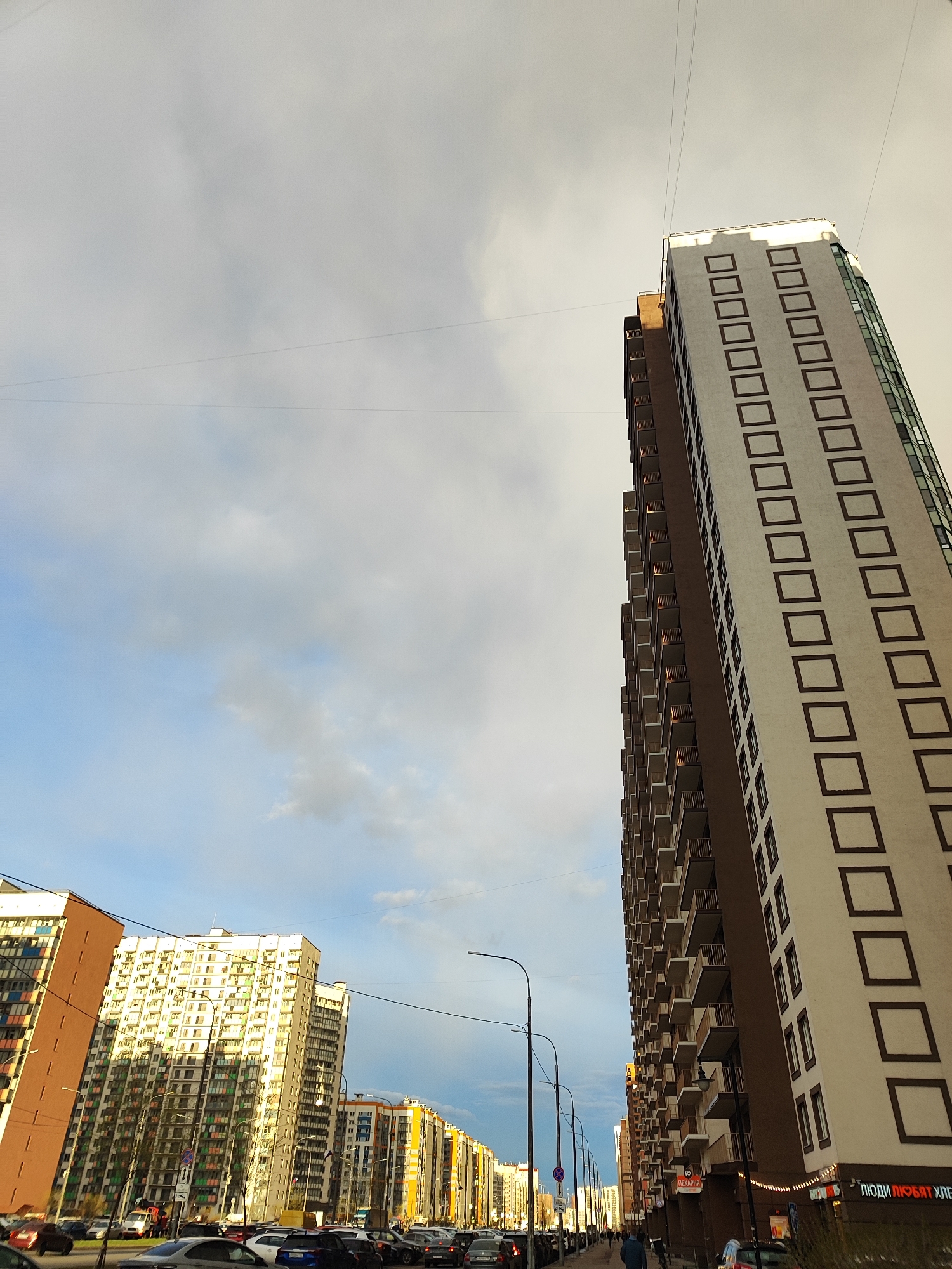 The image captures an urban street scene dominated by modern, tall residential buildings under a partly cloudy sky. On the right, a large, light beige building with a repeating pattern of dark brown square frames and multiple balconies extends upward, filling a significant portion of the frame. Its side is dark brown. On the left, another tall, light-colored building reflects sunlight, and further down the street, a long row of diverse residential buildings in varying colors, including orange, yellow, and white, line the thoroughfare.
The street below is densely packed with parked cars on both sides, suggesting a busy residential area. Several streetlights and overhead wires crisscross the view. A few pedestrians are visible walking on the sidewalks, indicating daily life. The sky is predominantly overcast with soft grey clouds, but patches of blue sky are visible, particularly towards the left and center. The quality of light, with some buildings illuminated by a low-angle sun, suggests either late afternoon or early morning.
Visible text on the ground floor of the large building on the right reads "ЛЮДИ ЛЮБЯТ ХЛЕБ" (PEOPLE LOVE BREAD). Further down the street, another sign indicates "ПЕКАРНЯ" (BAKERY), reinforcing the presence of food establishments in the area. The scene depicts a typical city environment, bustling with cars and residents.