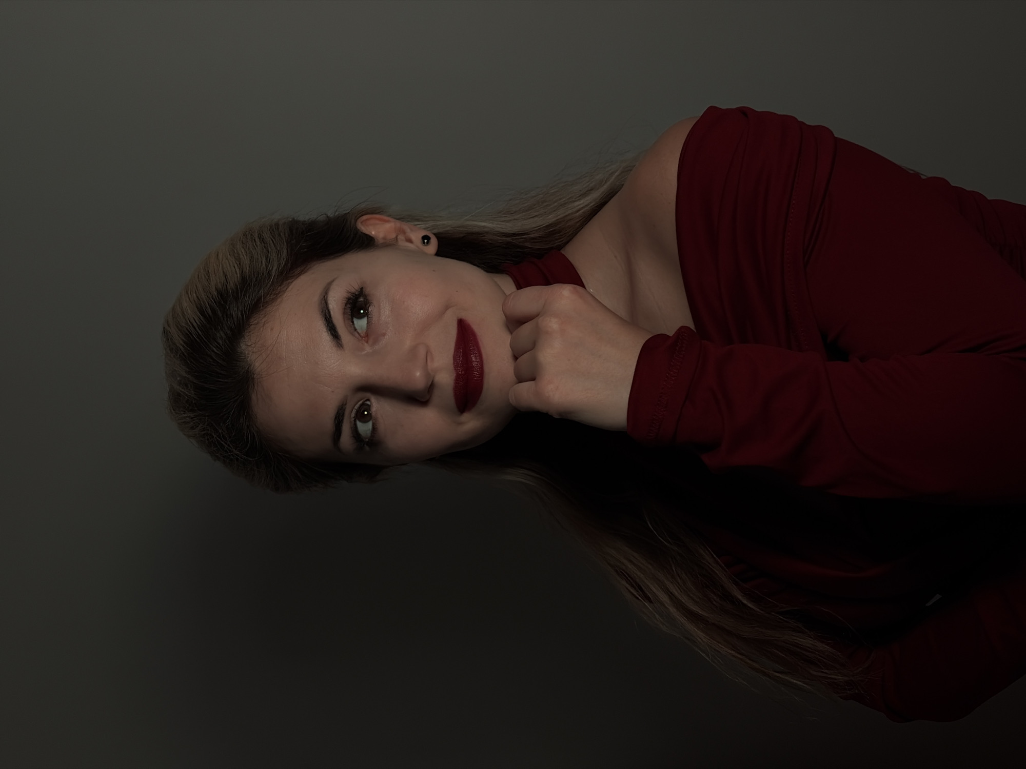 A woman with blonde hair and fair skin is shown from the chest up, angled to the left. She is wearing a long-sleeved, burgundy-colored top with an off-the-shoulder design. Her left hand is positioned near her chin. She has dark lipstick and noticeable eye makeup. Her hair is styled with some darker strands visible. The background is a solid, dark grey. No information about Budva, Montenegro can be extracted from the image.