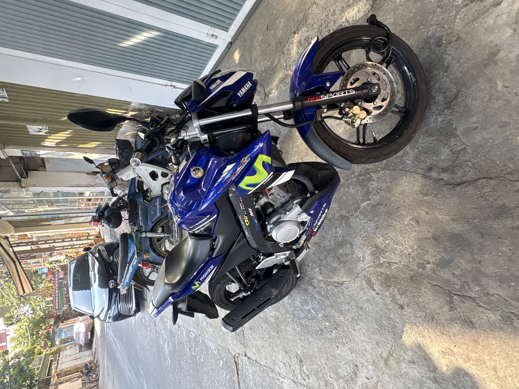 The image depicts a street scene in Nha Trang Ward, Vietnam, featuring a blue Yamaha motorcycle parked on the sidewalk. The motorcycle has prominent markings and logos: "Movistar," "brembo," "Revs Your Heart," "DELTABOX CRGC EDITION RACINGART," "YAMAHA," "SPORTS," and "YAMALUBE." It is parked alongside other motorcycles and a black Toyota car. In the background, shops and other buildings line the street, with trees and street signs visible. The scene is bathed in natural sunlight.