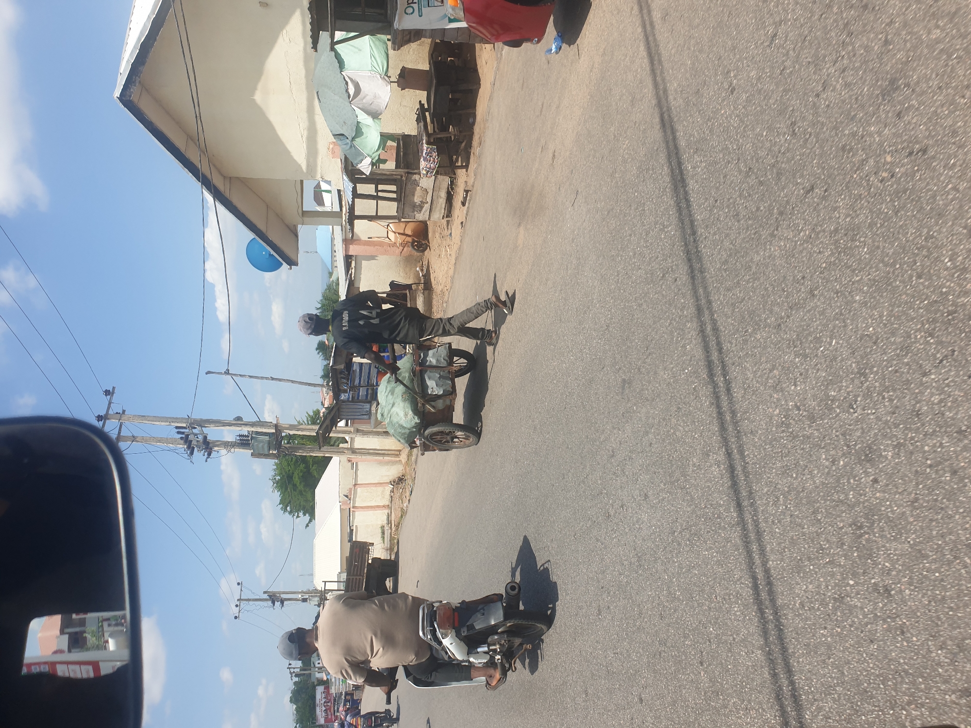 The image shows a street scene in Lokoja, Nigeria. The camera appears to be positioned from inside a vehicle. A man walks alongside a tricycle cart loaded with bulky items wrapped in green tarp. A second man is riding a motorcycle in the same direction. The street is paved and lined with buildings of varying construction. Utility poles are visible along the side of the road. The sky is blue with scattered clouds.