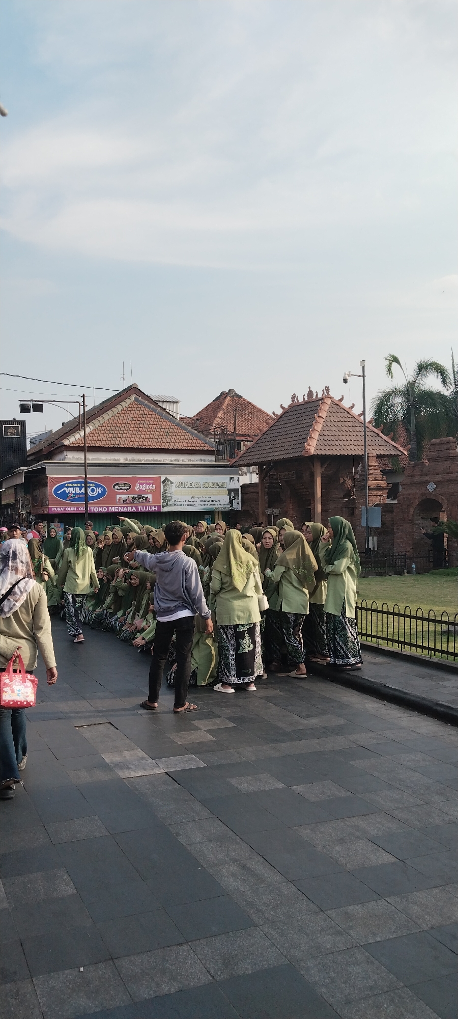 A group of young women in light green hijabs and patterned skirts are gathered in an outdoor area with a stone-tiled walkway. One woman in the foreground, on the left, is wearing a light gray hijab with a white pattern and is carrying a pink bag. Another person, facing away from the camera and towards the group of women, is wearing a gray hooded sweatshirt and black pants. This individual is gesturing with their right hand. The women are positioned in front of a building with an arched entryway and wooden latticework, which is adjacent to a brick wall. In the background, there are more buildings with tiled roofs and a palm tree. A black metal fence runs along the right side of the scene. Signs with text are visible on the building in the middle background, including one that reads "MUBAROK". The sky is partly cloudy and a bright blue. This scene appears to be located in the city of Kauman, Indonesia, likely a public space or tourist area.
