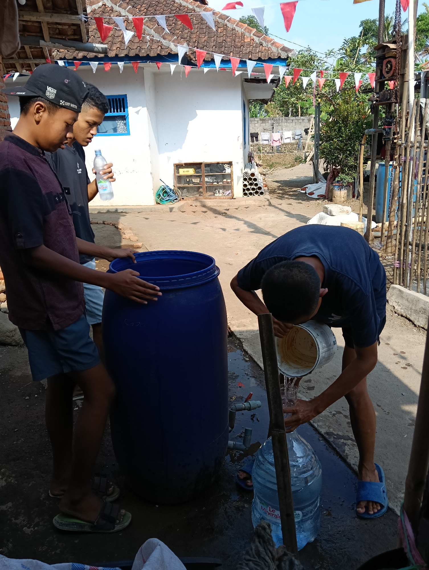 The image depicts three young males engaged in filling a large blue plastic drum and a large plastic water jug. The scene appears to be in a residential outdoor area, possibly a backyard or alleyway, in Tasikmalaya, Indonesia. In the background, a white building with a red-tiled roof is visible, adorned with red and white triangular flags. Various objects are scattered around, including PVC pipes, coiled hoses, and laundry hanging on a line. The ground is paved with concrete in some areas and dirt in others, with visible puddles of water.
The individuals are dressed casually. One young male on the left, wearing a maroon and black short-sleeved shirt and blue shorts, stands near the blue drum, with his hands resting on its rim. He is also wearing sandals. To his right, another young male, in a black t-shirt and blue shorts, holds a partially filled plastic water bottle. The third individual, on the right side of the frame, is actively pouring water from a white bucket into a large plastic water jug, using a bamboo pole to steady the jug. He is wearing a dark blue t-shirt and blue flip-flops. The action suggests a communal effort, potentially for water storage or distribution. The presence of the building and flags may indicate a local event or celebration.