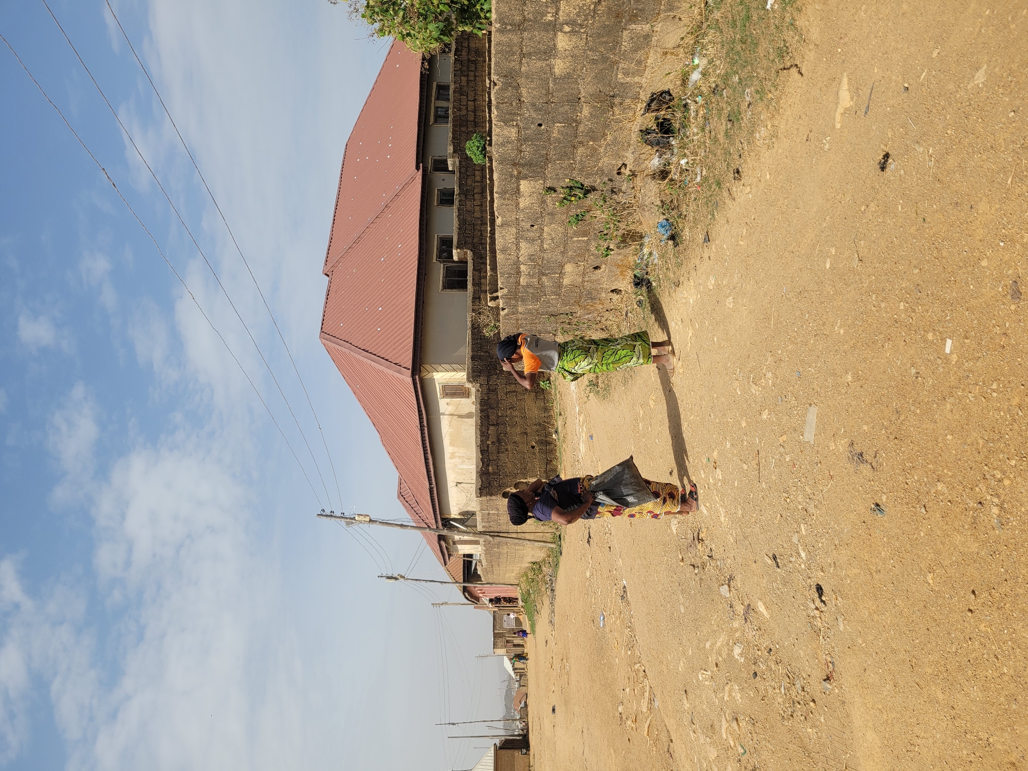 This image captures a scene in Barakin Akawo, Nigeria, depicting an unpaved dirt road flanked by buildings and utility poles under a clear, bright sky. The time of day appears to be late morning or early afternoon, with distinct shadows cast by subjects, suggesting bright sunlight.
Two women are visible on the road, engaged in everyday activities. In the foreground on the right, a woman stands facing away from the viewer, wearing a dark blue top and a vibrant, multi-colored patterned skirt or wrapper. She appears to be carrying a dark bag or item, possibly on her head or shoulder. Further back and slightly to the left, another woman is seen wearing an orange top and a green patterned skirt. She stands with her hands clasped or on her hips, looking towards the left side of the frame. Both women appear to be barefoot or wearing very simple footwear, indicative of the local environment.
To the left of the road, a prominent two-story building with a reddish-brown corrugated metal roof stands. Its walls are a mix of exposed concrete blocks and plastered sections, with several windows visible on the upper floor. Further down the road, more modest buildings and structures can be glimpsed in the distance. Wooden utility poles with multiple overhead wires line the left side of the road, receding into the background.
The road itself is a dusty, light brown dirt path, showing signs of wear and scattered debris. Along the right edge of the road, near the base of a low concrete block wall, there are visible piles of trash, including black plastic bags and other refuse, suggesting local waste management practices. Some dry grass and sparse vegetation grow along the roadside and near the buildings. The sky above is a pale blue with scattered wispy white clouds. The overall scene portrays a typical day in a bustling, semi-urban or rural community. No discernible text is visible in the image.