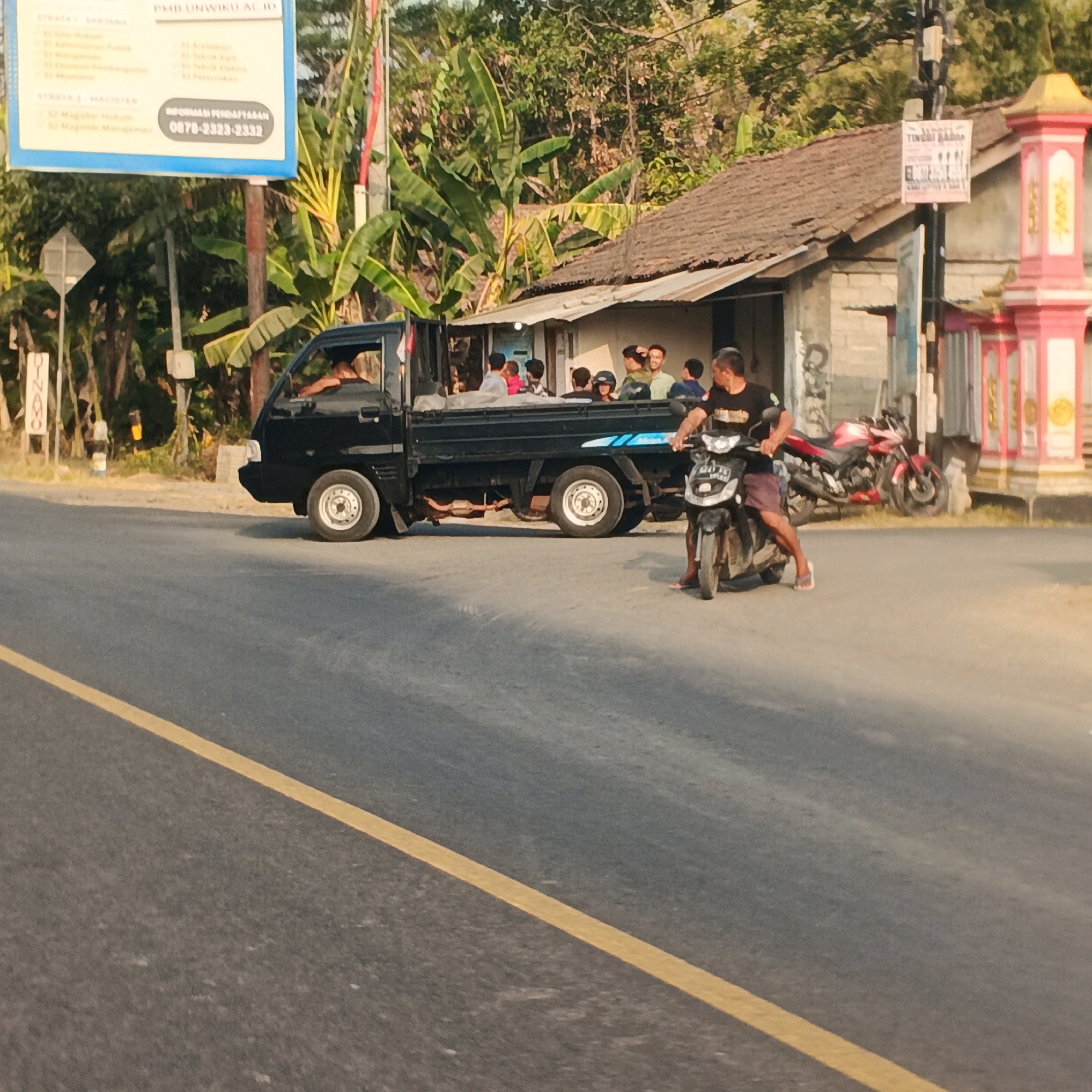 In Menganti, Indonesia, a black utility truck is parked on the side of a road. The truck bed is occupied by at least six individuals, including several young people and adults. The driver of the truck is visible in the cabin. In the foreground, a man is riding a motorcycle, looking towards the truck. Behind the motorcycle, a red motorcycle is parked. A residential building with a rustic roof is situated behind the truck and motorcycles, with lush green foliage surrounding the scene. A sign with Indonesian text is visible on a pole near the road. The road has a yellow line marking its edge. The lighting suggests it is either early morning or late afternoon.