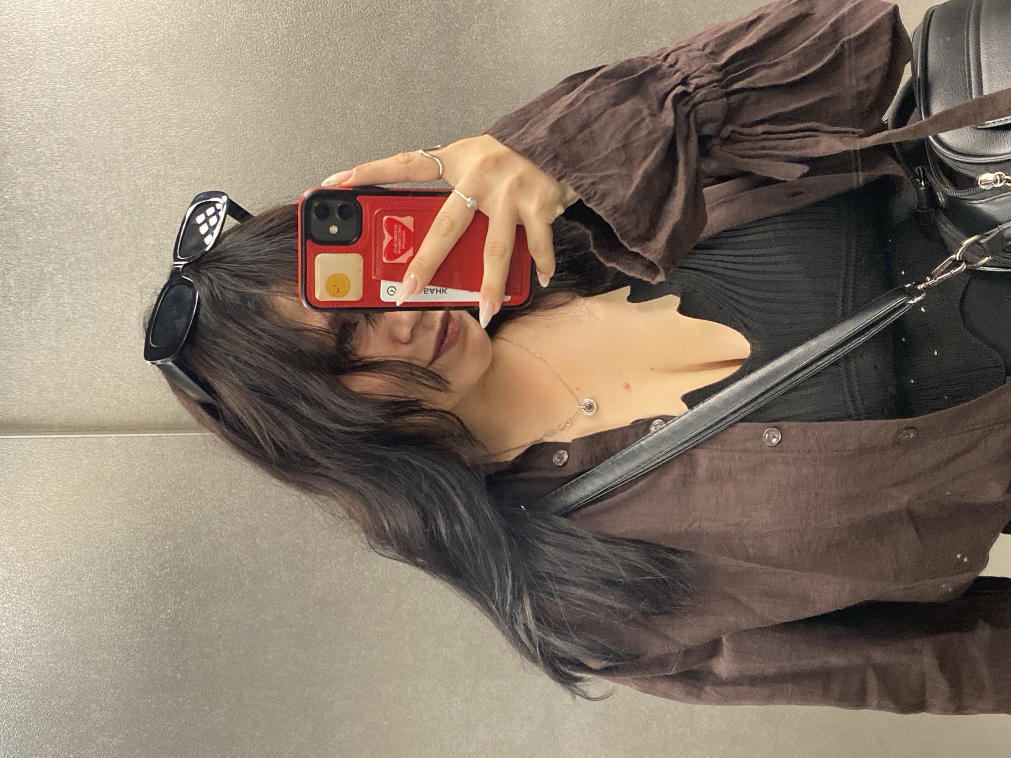 A young woman with dark, wavy hair, wearing a brown button-up shirt over a black ribbed top, takes a selfie in an elevator. She is holding a red phone with a sticker of a fried egg on the back. She is also wearing sunglasses on her head, a silver necklace, and two rings on her right hand. A black purse is slung across her shoulder. The background appears to be a brushed metal wall, typical of an elevator. The image is taken from the chest up.