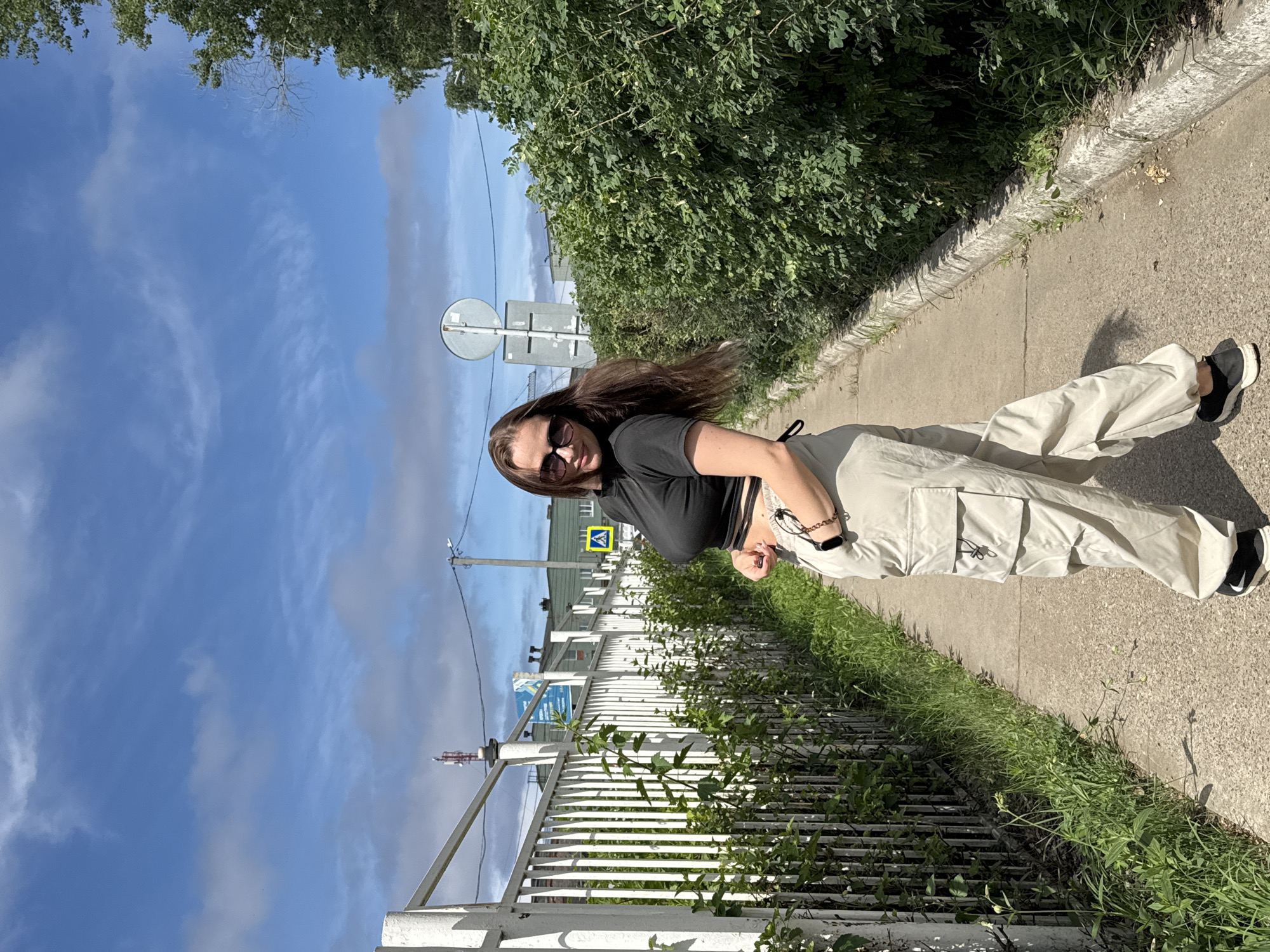 A young woman with long brown hair and sunglasses stands on a paved path in Chaikovsky, Russia. She is wearing a black crop top and light-colored cargo pants. To her left is a white metal fence bordering a grassy area with some bushes. To her right is a dense green hedge. In the background, there are buildings and a cell tower under a partly cloudy blue sky. A yellow pedestrian crossing sign is visible near the hedge.