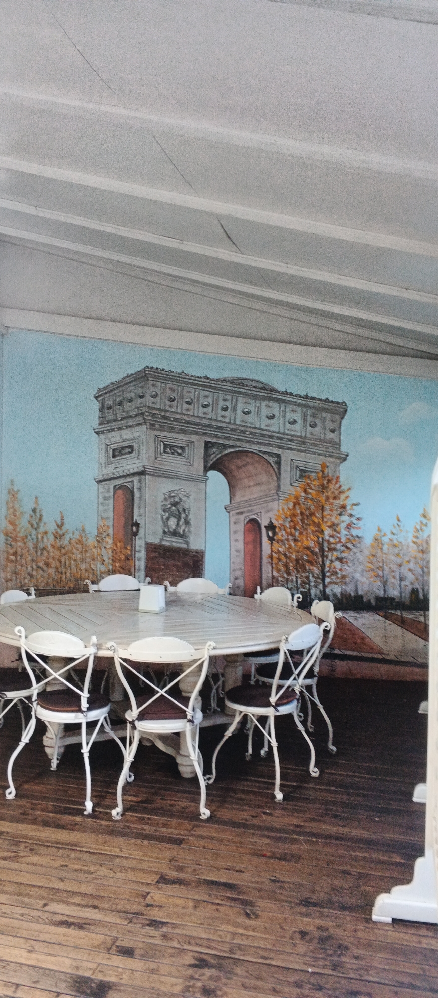 The image depicts a room with a mural on the wall. The mural features a depiction of the Arc de Triomphe, with trees and a blue sky in the background. In the foreground, a large oval table is surrounded by several white chairs with dark seats. A white napkin dispenser is placed in the center of the table. The floor is made of wooden planks. The overall impression is that of an indoor dining area designed with a Parisian theme. There are no people visible in the image. The provided information suggests this scene is located in Berehove, Ukraine.