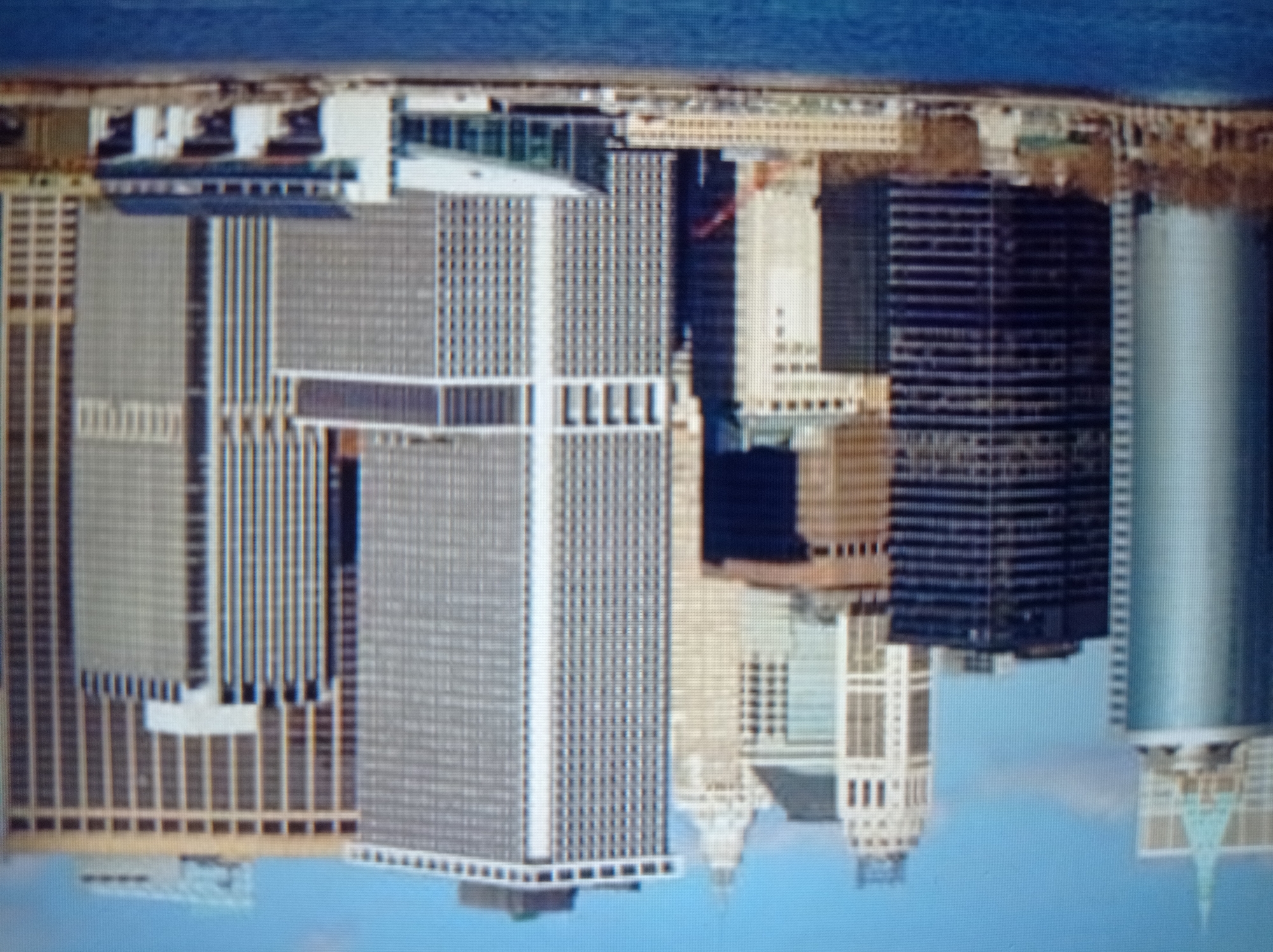 The image depicts a cityscape dominated by numerous tall buildings, suggesting an urban environment. In the foreground, a body of water, possibly a river or harbor, is visible. The buildings are a mix of architectural styles, with some featuring modern glass facades and others having more traditional designs with brick or stone exteriors. Several skyscrapers stand prominently, showcasing a dense concentration of high-rise structures. The sky above is clear blue with a few wispy clouds. The overall impression is that of a bustling metropolis. This scene is not in Mukachevo, Ukraine; it is likely New York City.
