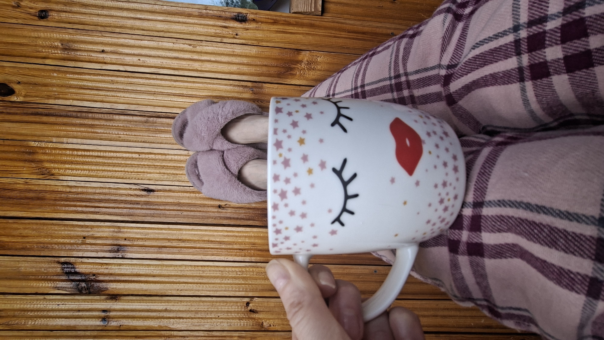 In Забалоцкі сельскі Савет, Belarus, a first-person, downward-angle photograph captures the lower body of an individual resting against a vertical wooden plank surface. The individual's legs are covered by pink and maroon plaid pajama pants. Their feet are encased in fuzzy, light purple slide slippers, with bare skin visible above the ankles. A left hand, with visible fingernails, holds a white ceramic mug featuring a graphic of closed black eyelashes, red lips, and scattered pink and gold stars. The background is composed of natural light brown wooden planks, exhibiting visible grain and several dark knots.