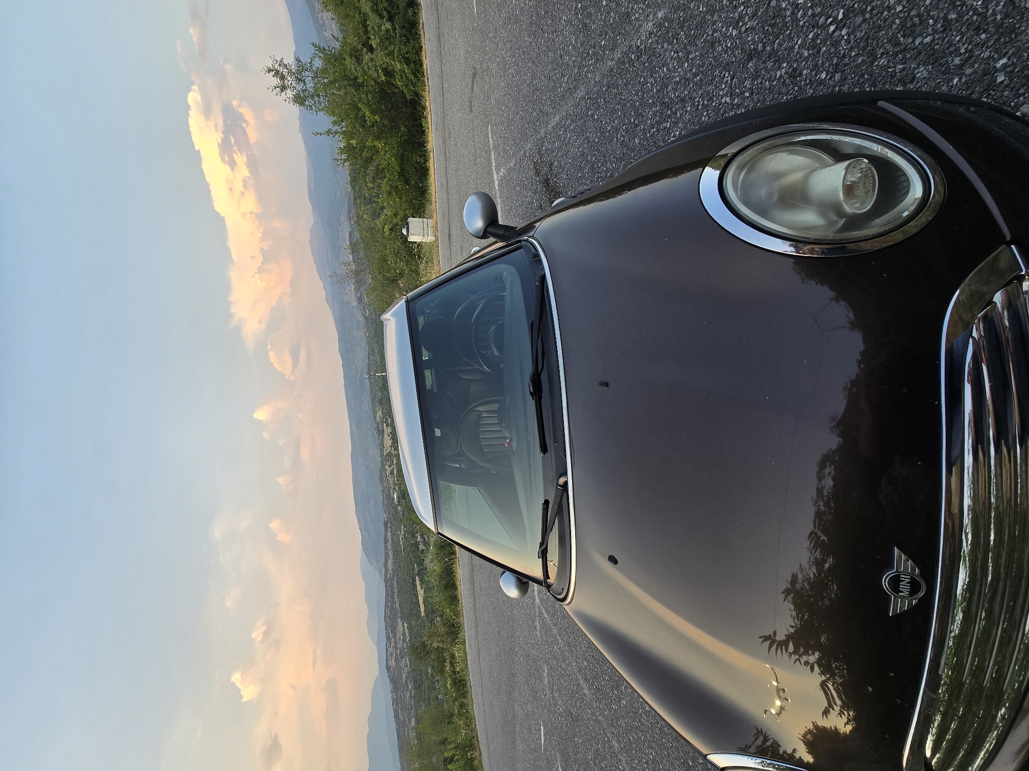 A dark brown Mini Cooper is parked on the side of a paved road, facing the camera. The car's hood is prominently featured, with reflections of trees and the sky visible on its surface. The Mini Cooper logo is centered on the hood. The car has a silver roof and silver side mirrors. The driver's side headlight, a large circular lamp surrounded by a chrome trim, is visible in the lower right portion of the image.

The car is positioned on a slight curve of the road, with the pavement extending into the distance. To the right of the road, there is a grassy shoulder and a white concrete marker. Beyond that, a dense line of green trees forms a backdrop.

In the background, a vast, hilly landscape unfolds under a sky filled with soft, pastel-colored clouds, suggesting either sunrise or sunset. The distant mountains are hazy and blue, fading into the horizon. The overall impression is a scenic, possibly rural or mountainous location. The location is in Medun, Montenegro.