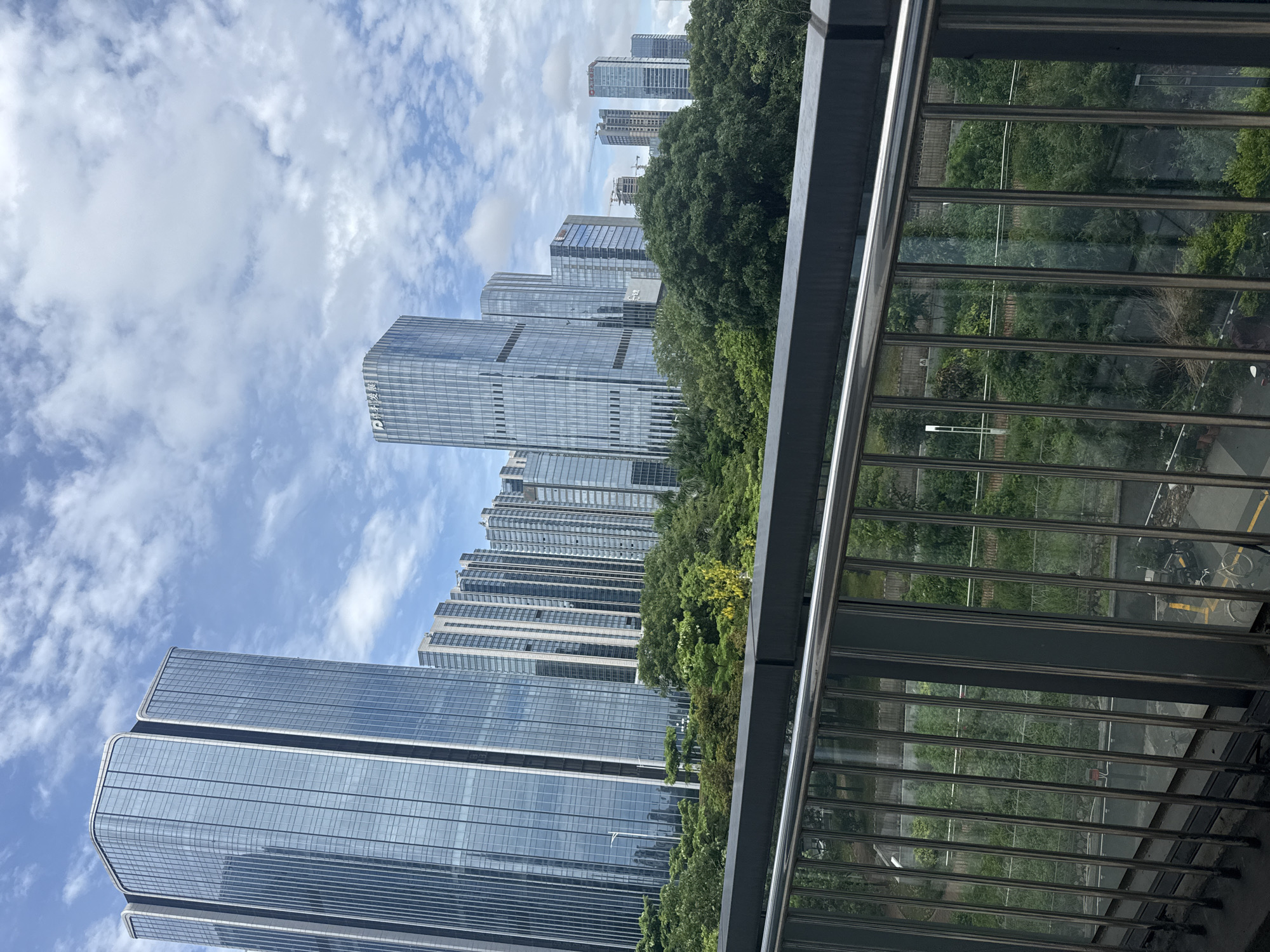 This image captures a view of modern skyscrapers in Tianhe District, China, under a partly cloudy sky. In the foreground, a metal railing from an elevated walkway or bridge runs horizontally across the frame, with vertical metal bars below. Beyond the railing, lush green trees create a dense canopy. In the mid-ground, various modern office buildings with glass facades rise majestically, some reflecting the sky and surrounding environment. A few of these buildings exhibit distinctive architectural designs with rounded corners and vertical lines. The background is dominated by the sky with scattered white clouds. The overall scene depicts a blend of urban development and natural greenery within a city. No people are visible in the image.