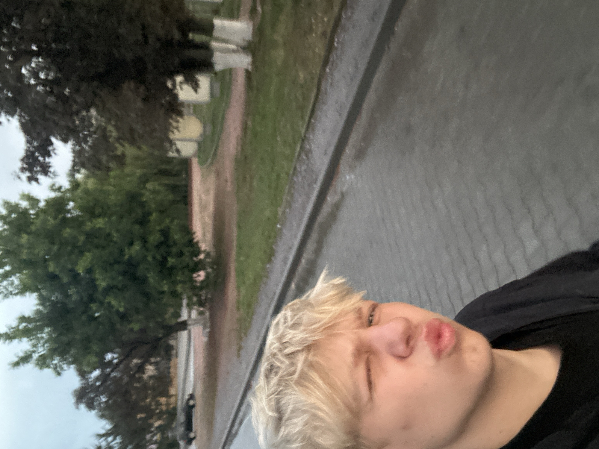A close-up selfie shows a young person with blonde hair puckering their lips and winking. They are wearing a black t-shirt and a black hooded garment. The background suggests an outdoor setting in Khmilnyk, Ukraine, with trees, a paved walkway, and a street visible. A dark-colored car is parked in the distance on the left side of the image, and several light-colored structures resembling benches or planters are present along a grassy area on the right. The overall lighting indicates it is likely late afternoon or early evening.