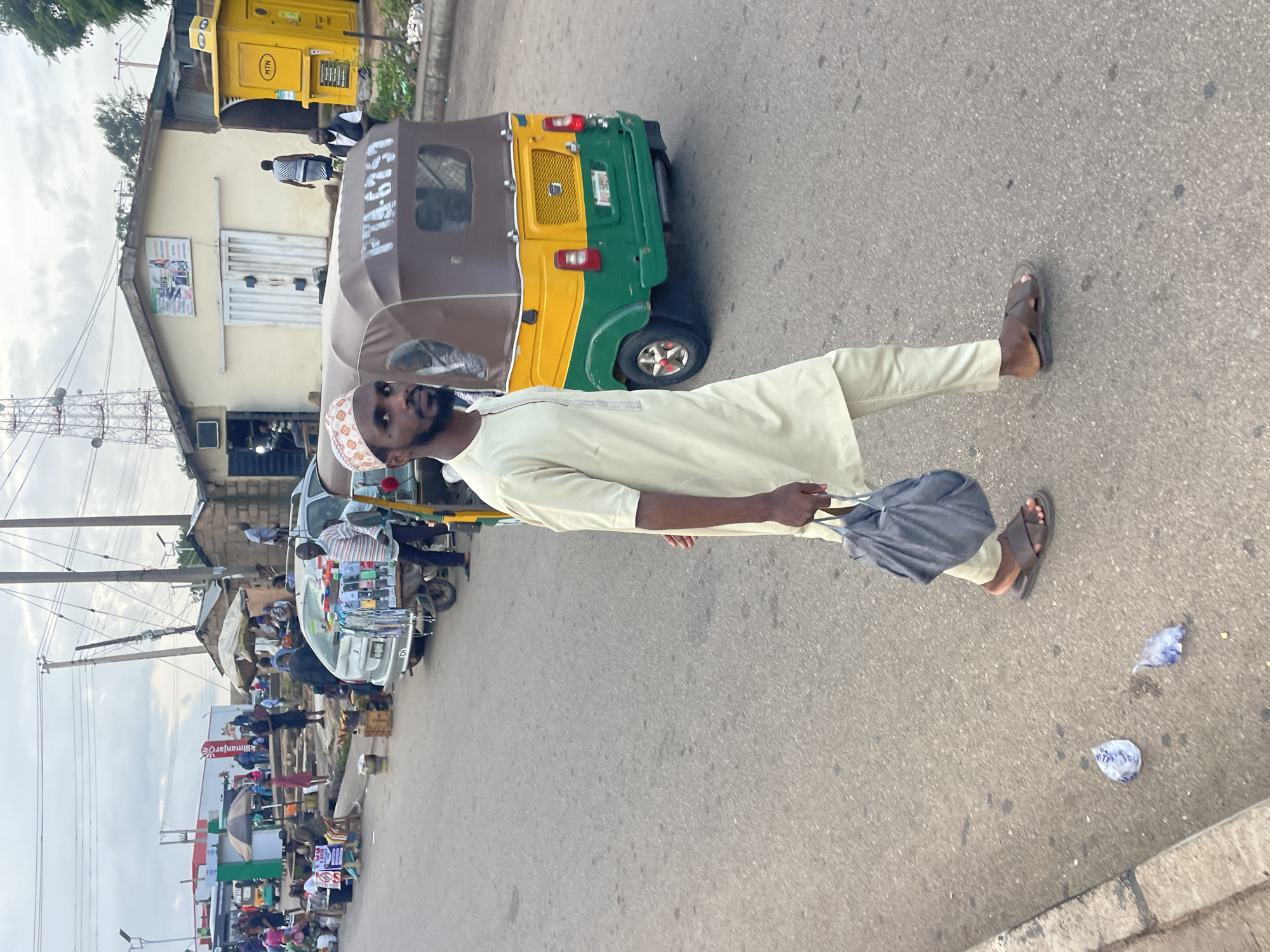 In Barakin Lerawi, Nigeria, a man in a pale yellow traditional outfit and a patterned cap walks on a paved street. He carries a dark gray bag in his right hand. He wears dark brown sandals. Behind him, a yellow and green auto-rickshaw with license plate PTA-6757 is parked. The street is lined with small shops and stalls. In the background, there are utility poles and a large radio tower. The sky is overcast.