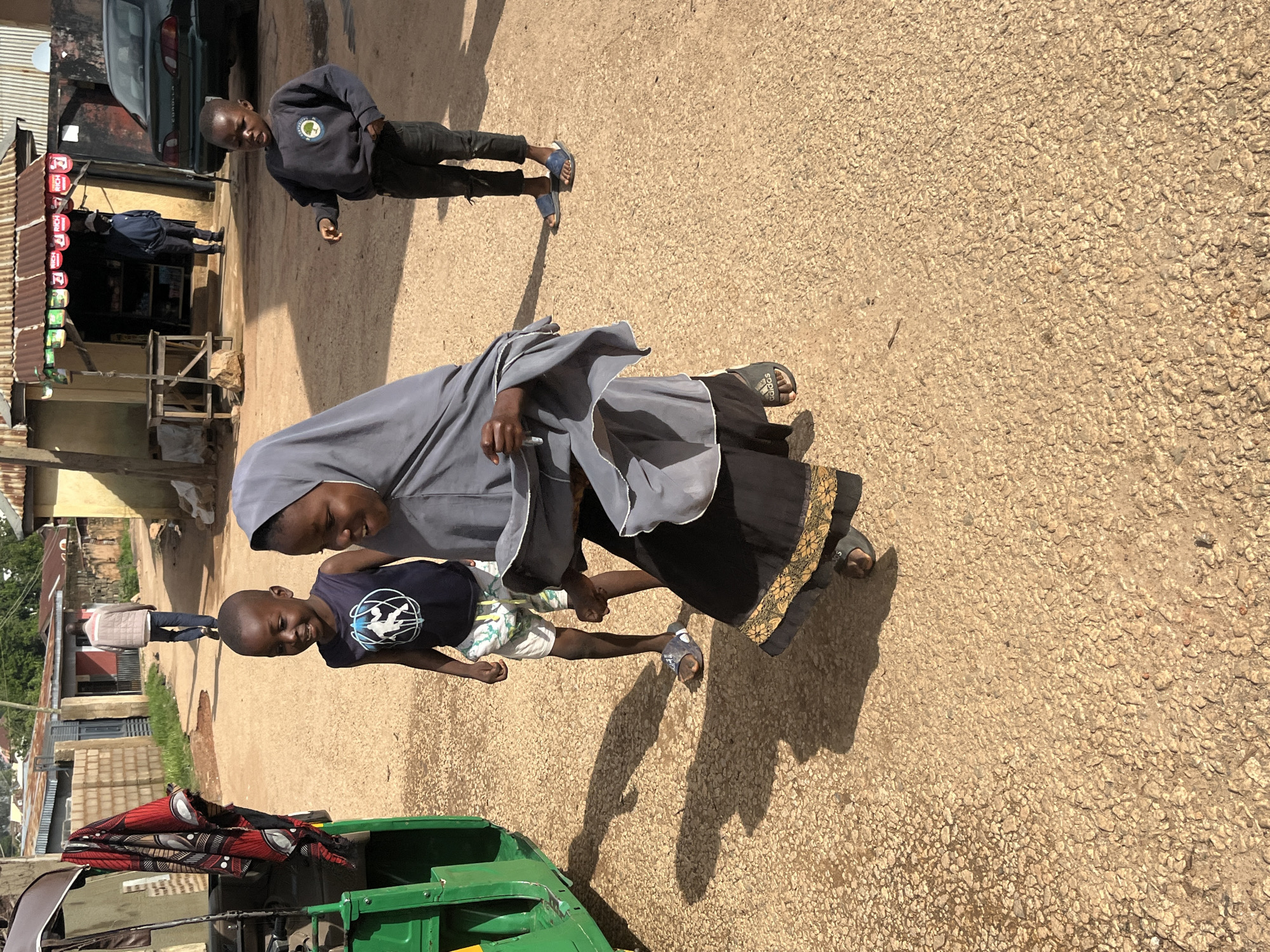 The image, taken in Jos, Nigeria, depicts a street scene under bright daylight, casting long shadows, suggesting it is either morning or late afternoon. The primary subjects are two individuals, an adult and a child, with another person visible in the background.
The adult, wearing a grey hijab and a black dress with a decorative gold border, is bending down, interacting with the child. The child, a young boy, is looking up at the adult with a wide smile, appearing joyful. He is dressed in a dark blue t-shirt with a white graphic and patterned shorts.
In the background, a man in a dark blue jacket and dark pants is walking away from the camera, towards a small shop with a corrugated metal roof. The shop appears to be stocked with various items displayed on shelves. Adjacent to the shop is a dark-colored vehicle.
In the foreground, part of a green auto-rickshaw, commonly known as a "Keke Napep," is visible. It is equipped with a colorful fabric covering, red and black patterned. The ground is a rough, sandy texture, typical of many roads in Nigeria. The overall atmosphere seems relaxed and everyday.