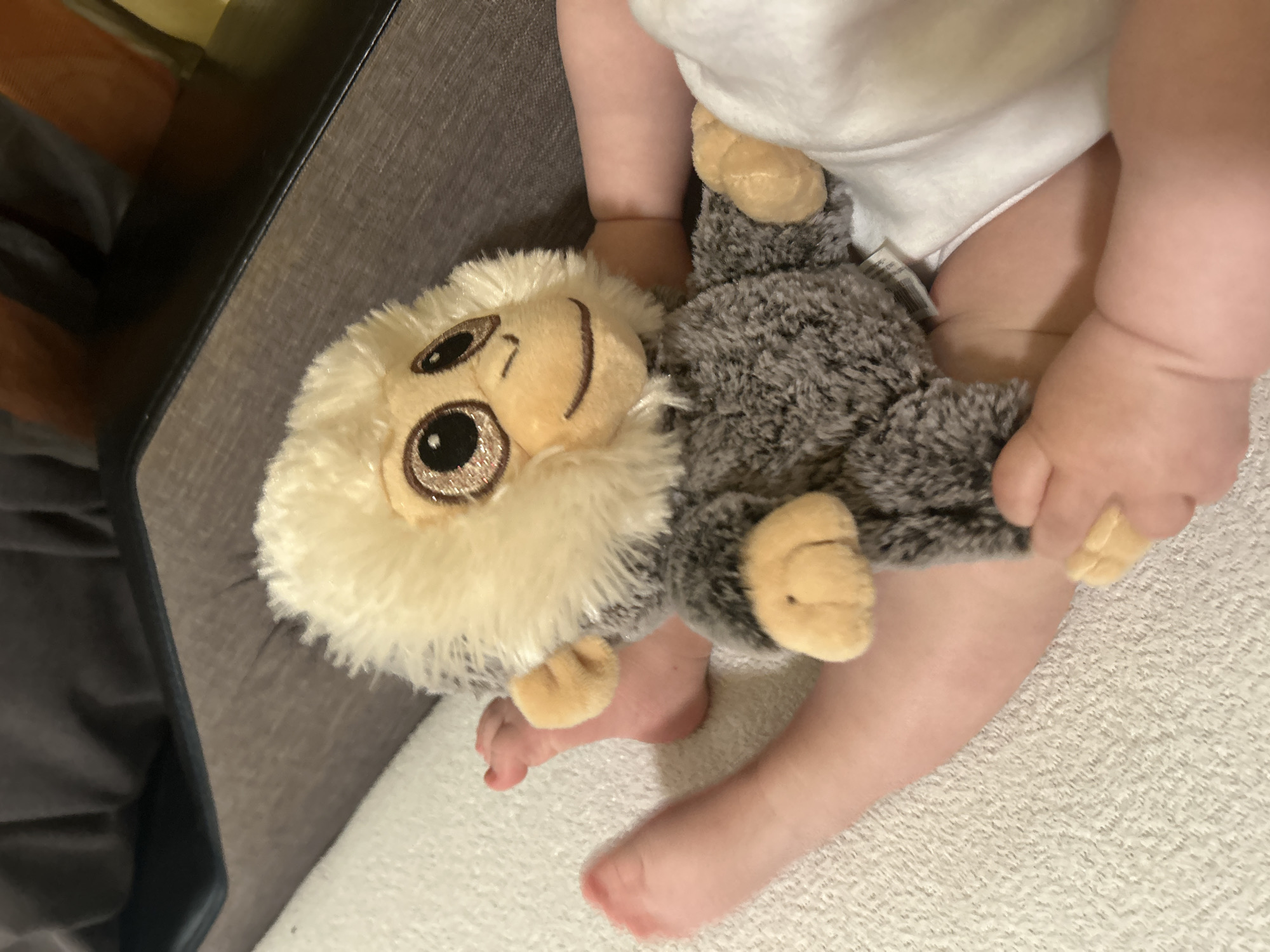 A close-up shot shows a baby lying on a white textured surface, possibly a bed or blanket. The baby is wearing a white bodysuit. The baby's feet are visible at the bottom of the frame. The baby is holding a plush monkey toy with a white fluffy mane, a tan face, and gray fur. The location is impossible to determine.
