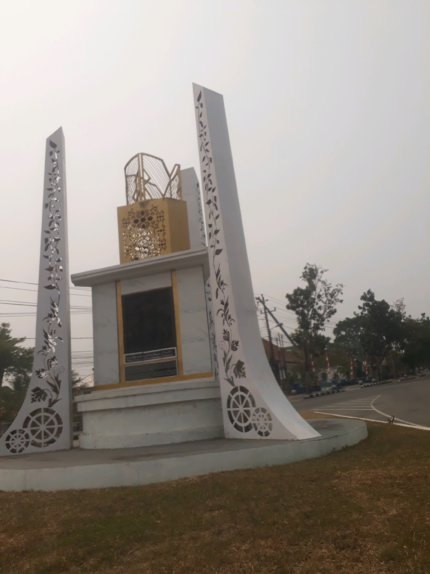 This is an outdoor, eye-level, medium shot of the Bundaran Tiga, a roundabout monument located in Pangkalpinang, Indonesia. The monument is white with gold accents and features intricate floral and circular cut-out designs on its tall, triangular side pillars. The central part of the monument has a gold, cage-like structure on top of a white box with a dark rectangular plaque. The sky is overcast and hazy. In the background, there is a road, trees, and power lines. The ground in front of the monument is covered in dry grass.