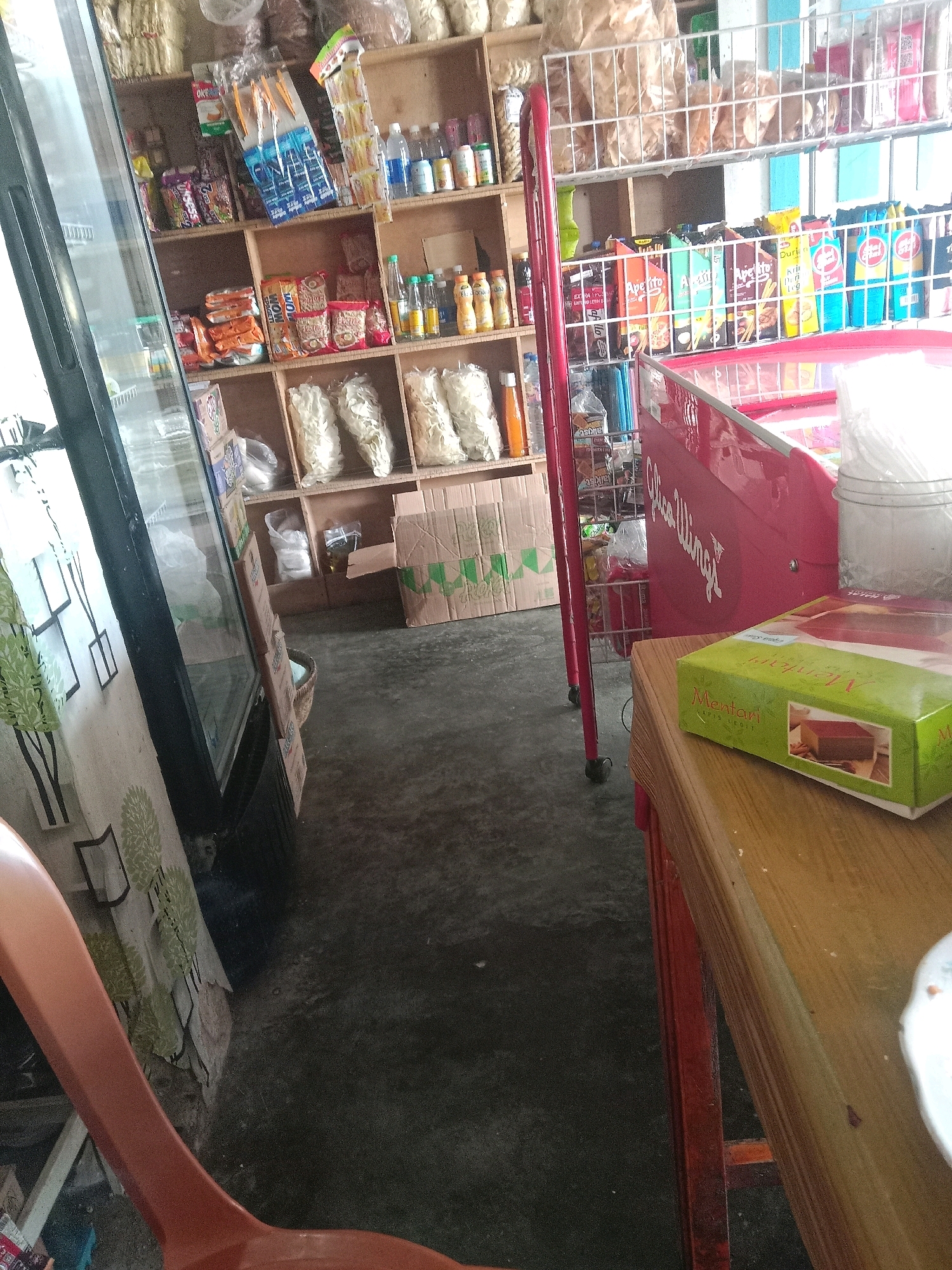 The image depicts a small convenience store, likely a warung, located in Kertek, Indonesia. The store is filled with various packaged food items and beverages displayed on wooden shelves and wire racks. Prominent items include snacks, candies, bottled drinks, and what appears to be pre-packaged bread or cakes. A red refrigerated display case is visible, along with a portion of a wooden counter. The floor is made of concrete. The overall impression is of a typical, modestly stocked Indonesian convenience store.